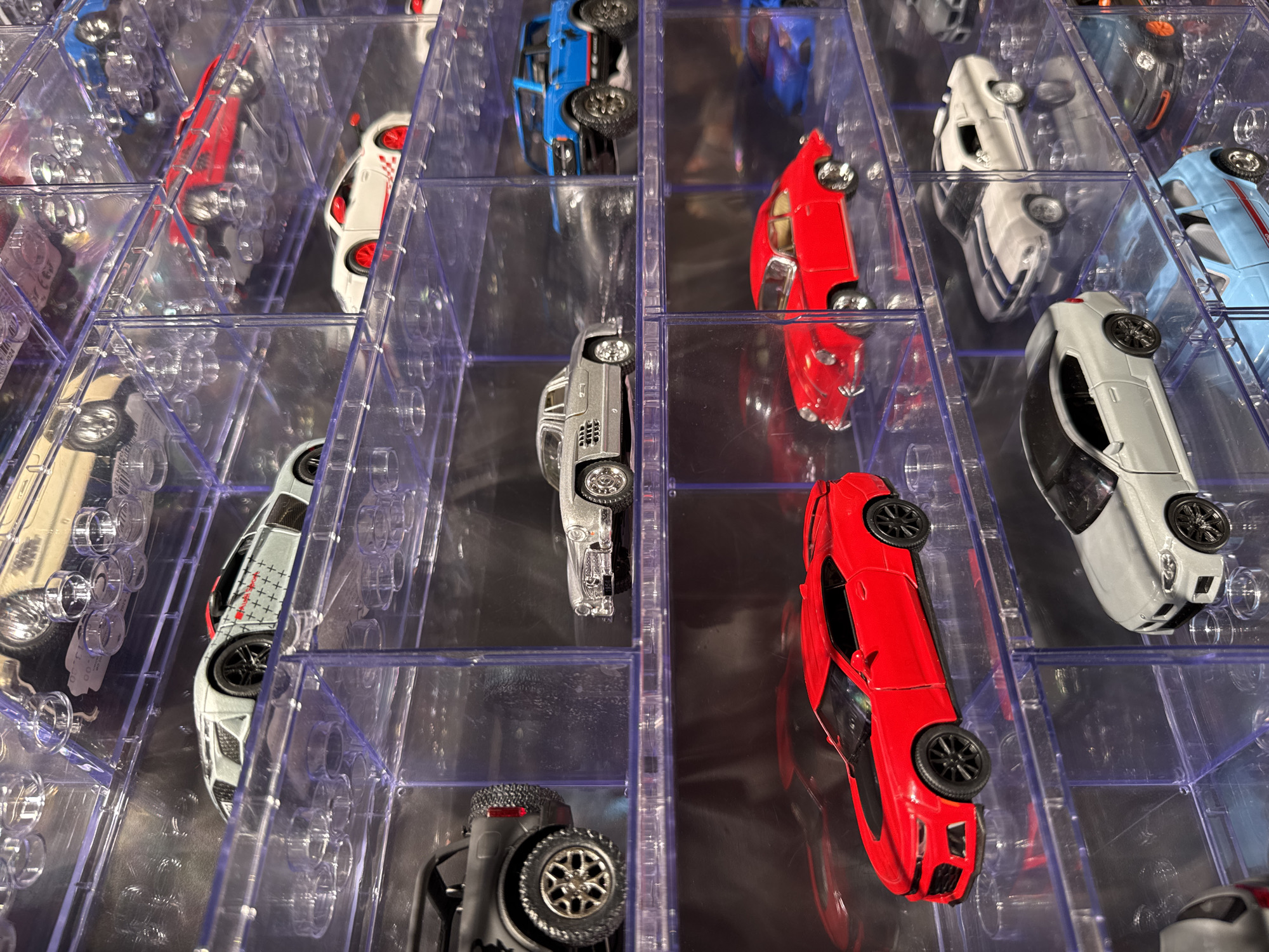The image displays a collection of miniature cars housed in clear plastic display cases. The cars are arranged in multiple tiers, showcasing a variety of makes and models, including sports cars, SUVs, and classic vehicles. The lighting appears to be artificial, illuminating the details of each toy car. The background is dark and out of focus, emphasizing the collection. There is no indication of the city of Otopeni, Romania, in the image itself or in any context provided with the image.