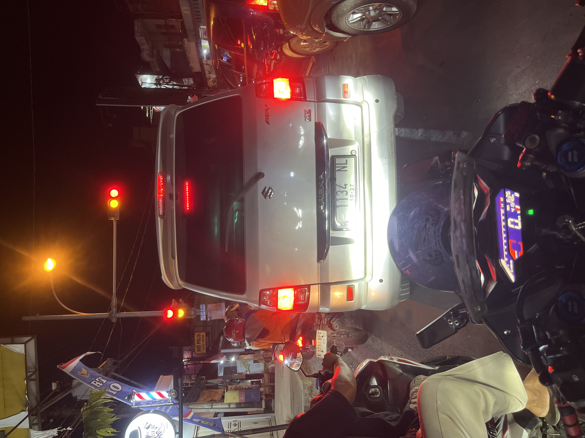 The image captures a night scene in Kalinyamatan, Indonesia, likely during a traffic jam. The perspective is from a motorcycle, looking forward at a silver Suzuki APV van. The van's brake lights and reverse lights are illuminated. To the left of the van, a motorcycle with a rider wearing an orange jacket and a red helmet is visible, along with a portion of another motorcycle in the foreground. Traffic lights are red and yellow, indicating a stop or caution. In the background, there are buildings, streetlights, and other vehicles. A barbershop sign and a police sign ("POS POLISI LALU LINTAS") are also discernible. The overall atmosphere is dark, with artificial lighting from vehicles and street lamps.