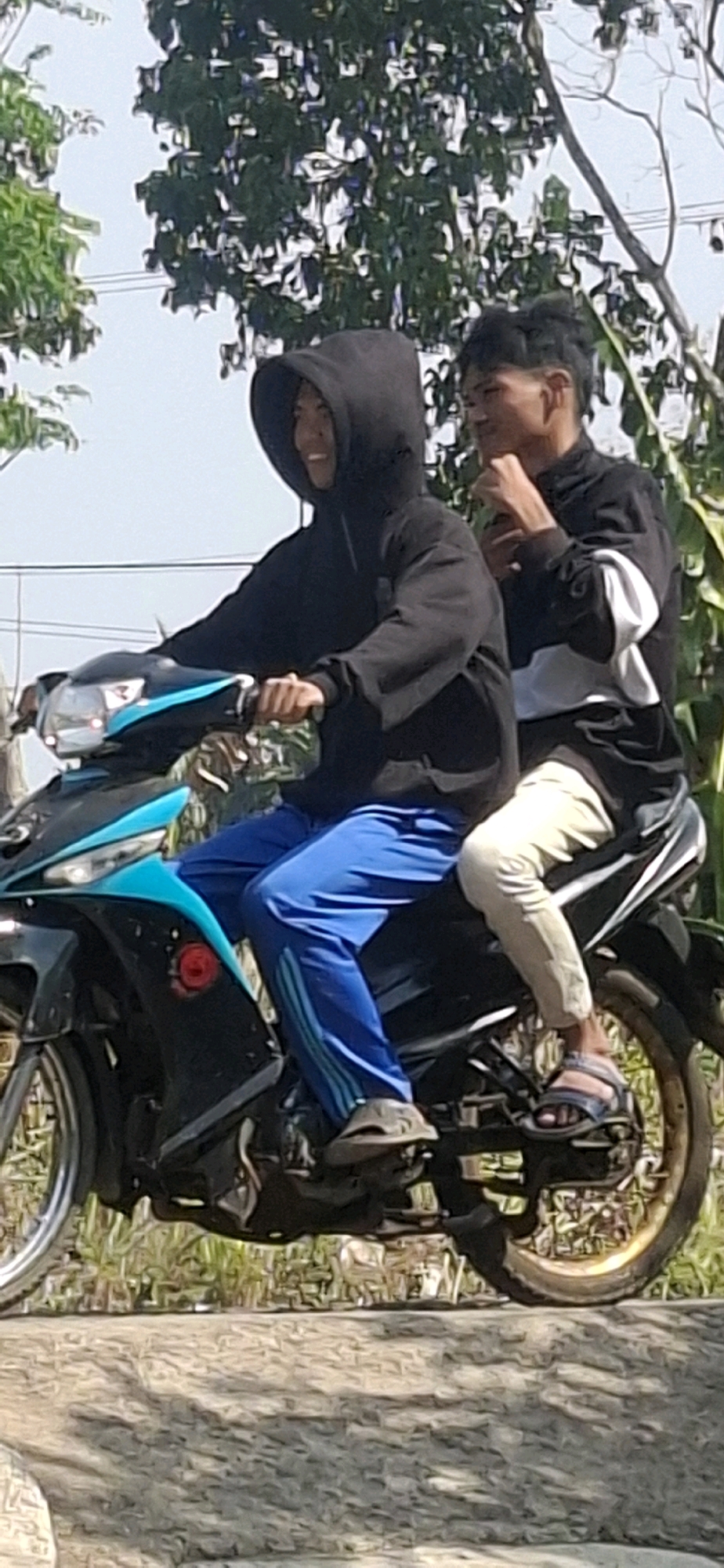 Two young males are riding a black and blue motorcycle. The rider in front, wearing a black hooded sweatshirt and blue athletic pants, is looking to the left and smiling. The passenger behind him, wearing a black jacket with white and gray stripes and light-colored pants, is looking to his right and appears to be touching his chin. They are on a paved surface, possibly a road, with vegetation and trees in the background. The sky is visible, suggesting it is daytime. The overall scene suggests a casual outing or transportation in a tropical or subtropical environment. While specific landmarks for Cibinong Hilir, Indonesia are not visible, the presence of lush greenery and overhead power lines are consistent with semi-urban or rural Indonesian settings.