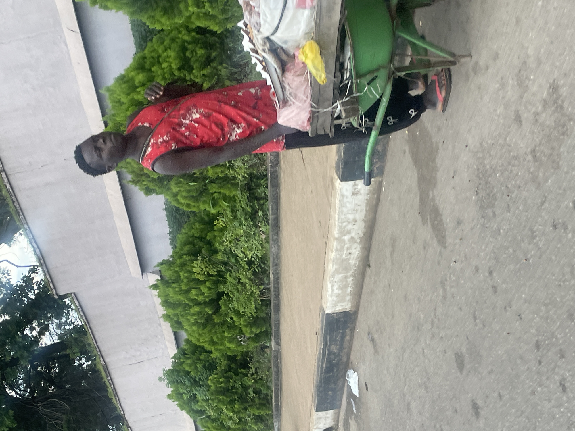 A young Black male, appearing to be in his late teens or early twenties, stands next to a green wheelbarrow loaded with various items, possibly goods for sale. He wears a red sleeveless shirt with a white, abstract pattern and black pants with a white symbol on the left leg. His right foot is adorned with a red flip-flop. The wheelbarrow is positioned on a paved surface, adjacent to a curb with black and white markings. Behind the curb, there is a patch of dirt or sand, and further back, a row of manicured green shrubs lines a light-colored, textured wall. Trees with dark foliage are visible at the top of the image, with some branches extending over the wall. The scene appears to be outdoors, in what could be a public space within Jos, Nigeria, given the context. The lighting suggests it is daytime. The wheelbarrow contains a pink plastic bag, a yellow plastic bag, and other items wrapped in white plastic or cloth. The man's gaze is directed to his right, and his left hand is resting on the wheelbarrow. A thin strap is visible across his chest, originating from the left side of his shirt. The overall impression is of a vendor or transporter preparing to move or display their wares.