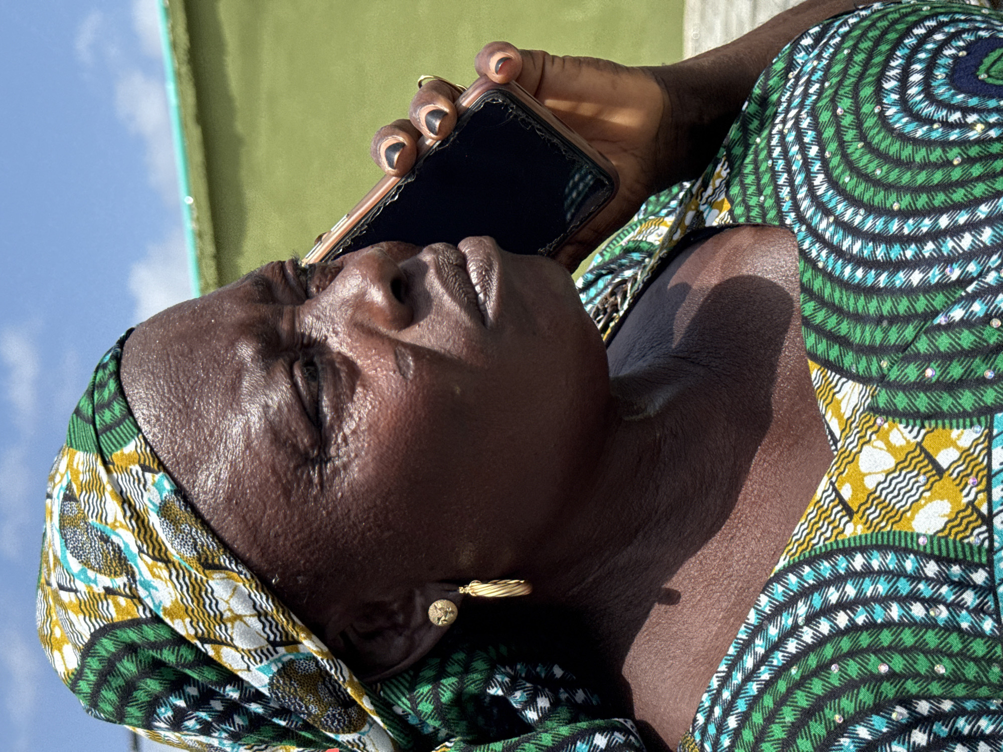 A close-up, angled shot shows a middle-aged, dark-skinned woman holding a damaged smartphone to her ear. She is outdoors on a partly sunny day, near a pale green and white structure. The woman is wearing a green, white, and yellow patterned headscarf and a matching top. She has dark fingernails and wears gold earrings. The scene is presumed to be in Ado, Nigeria.