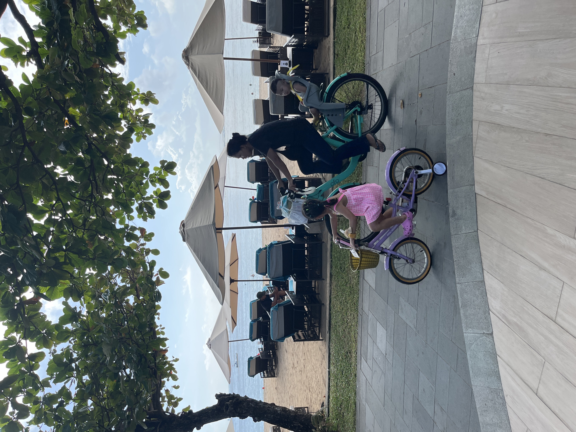 In Kuta Selatan, Indonesia, a woman rides a teal bicycle with a baby in a gray child seat attached to the rear. A young girl in a pink dress rides a purple bicycle in front of her. The bicycles are on a paved walkway. In the background, lounge chairs and umbrellas are set up on a sandy beach overlooking the ocean. Green trees with large leaves overhang the scene from the left side of the frame. The sky is blue with scattered clouds.