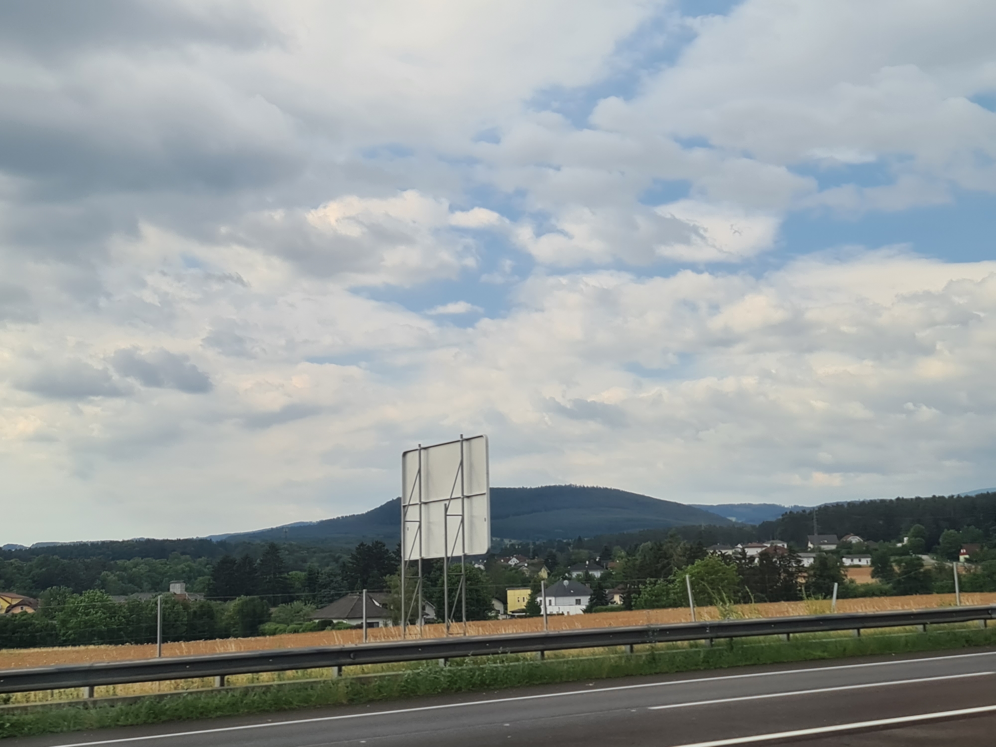 The image captures a scenic landscape viewed from a highway in Ramplach, Austria. In the foreground, a multi-lane road is visible, with a metal guardrail separating it from a golden field of crops. Beyond the field, a small village is nestled amongst rolling hills covered with lush green trees. In the middle ground, a large, blank billboard stands prominently on a metal frame. The background is dominated by a vast expanse of sky filled with a mix of white and gray clouds, with patches of blue peeking through. The overall scene suggests a rural setting with a hint of infrastructure from the highway and billboard.