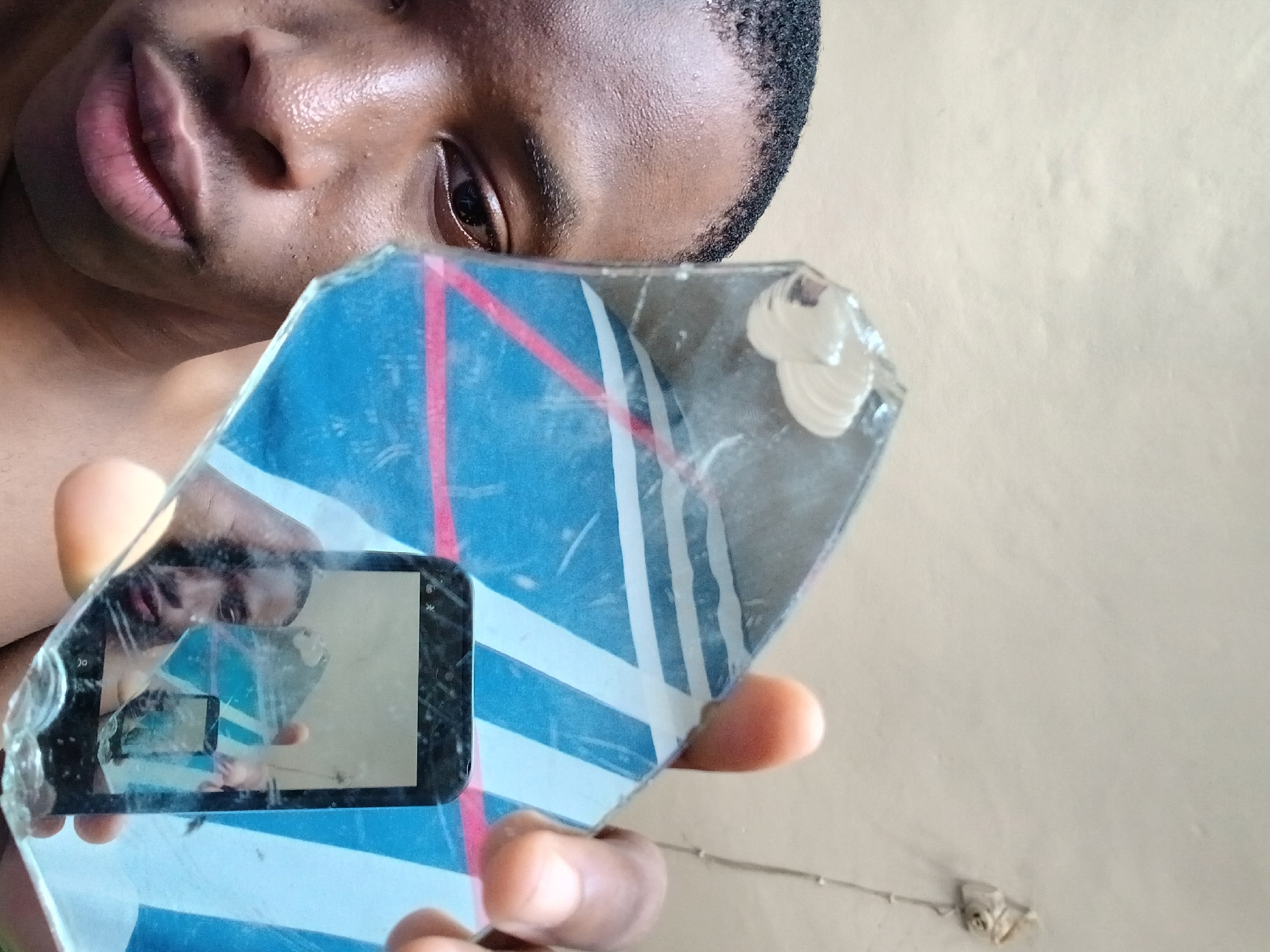A dark-skinned person is holding a piece of broken mirror, with a reflection of a phone screen capturing a reflection within the larger mirror piece. The mirror piece has a blue and white pattern with a red line and is cracked. The person's face is partially visible, including lips, nose, and one eye. The background appears to be a plain, light-colored wall. The location in Lagos, Nigeria cannot be determined.