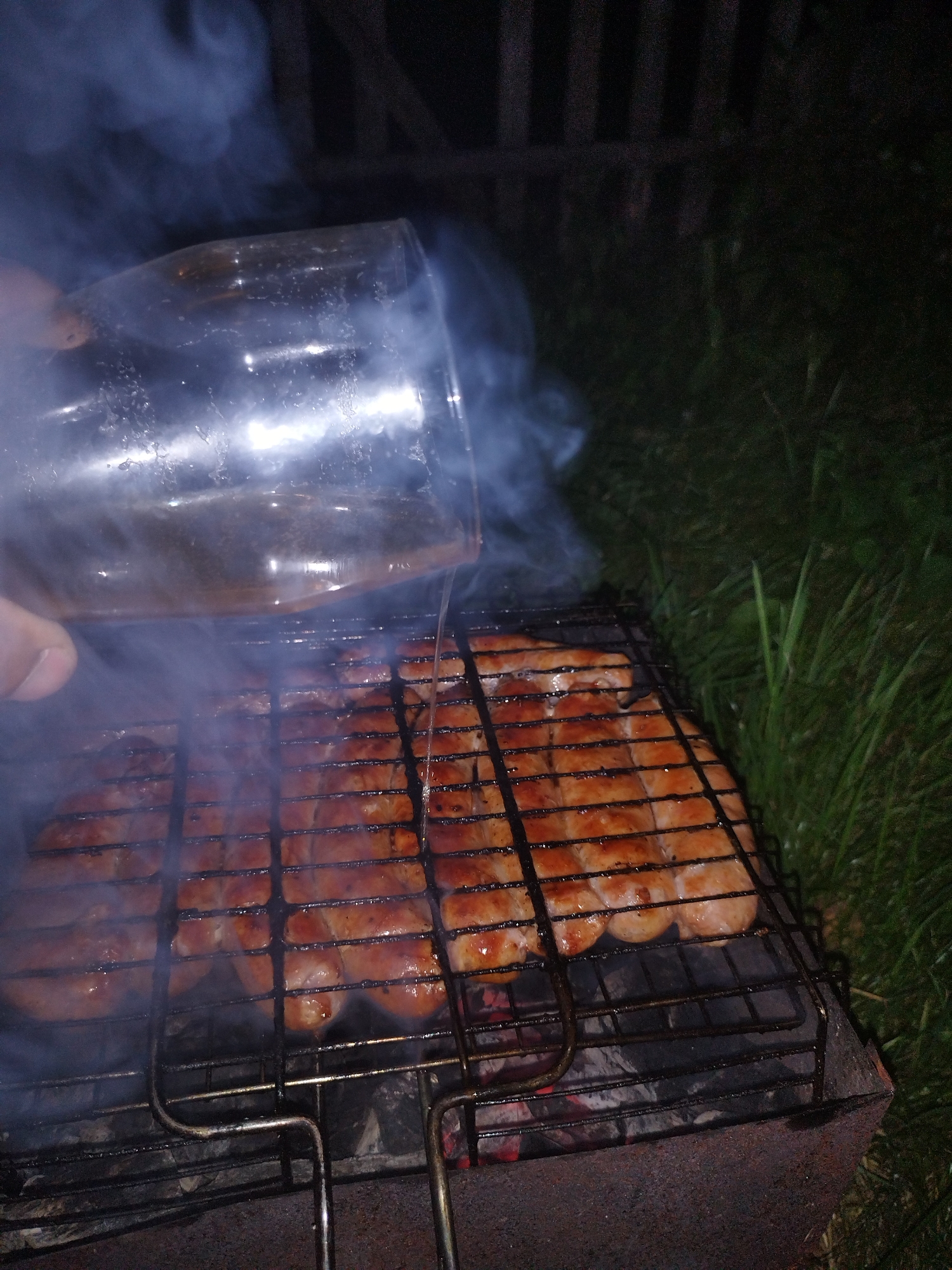 An outdoor grilling scene at night. The focus is on a grill rack filled with several sausages cooking over glowing embers. A hand is holding a clear glass, from which liquid is being poured onto the sausages, creating steam. The background is dark with some out-of-focus grass and a wooden fence visible. The scene appears to be in a backyard or park setting.