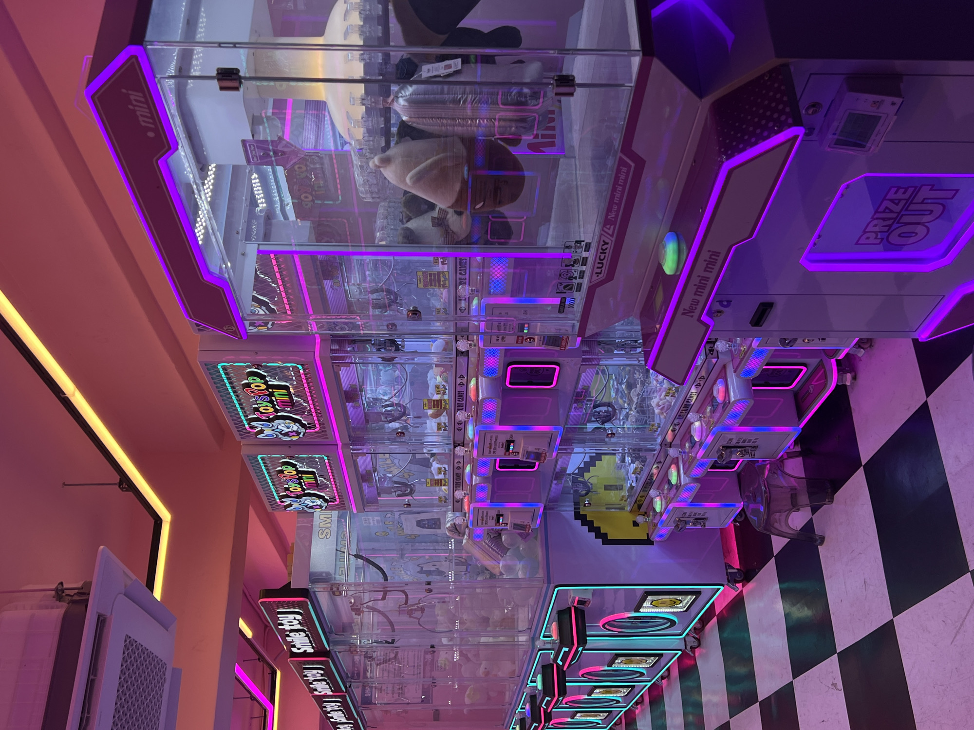 The image shows a row of claw crane arcade game machines, likely located in Seoul, South Korea, based on the lettering on the machines. The machines feature colorful neon lights, including purple, blue, and pink. The games are filled with various plush toys and prizes. The floor has a checkered black and white pattern.