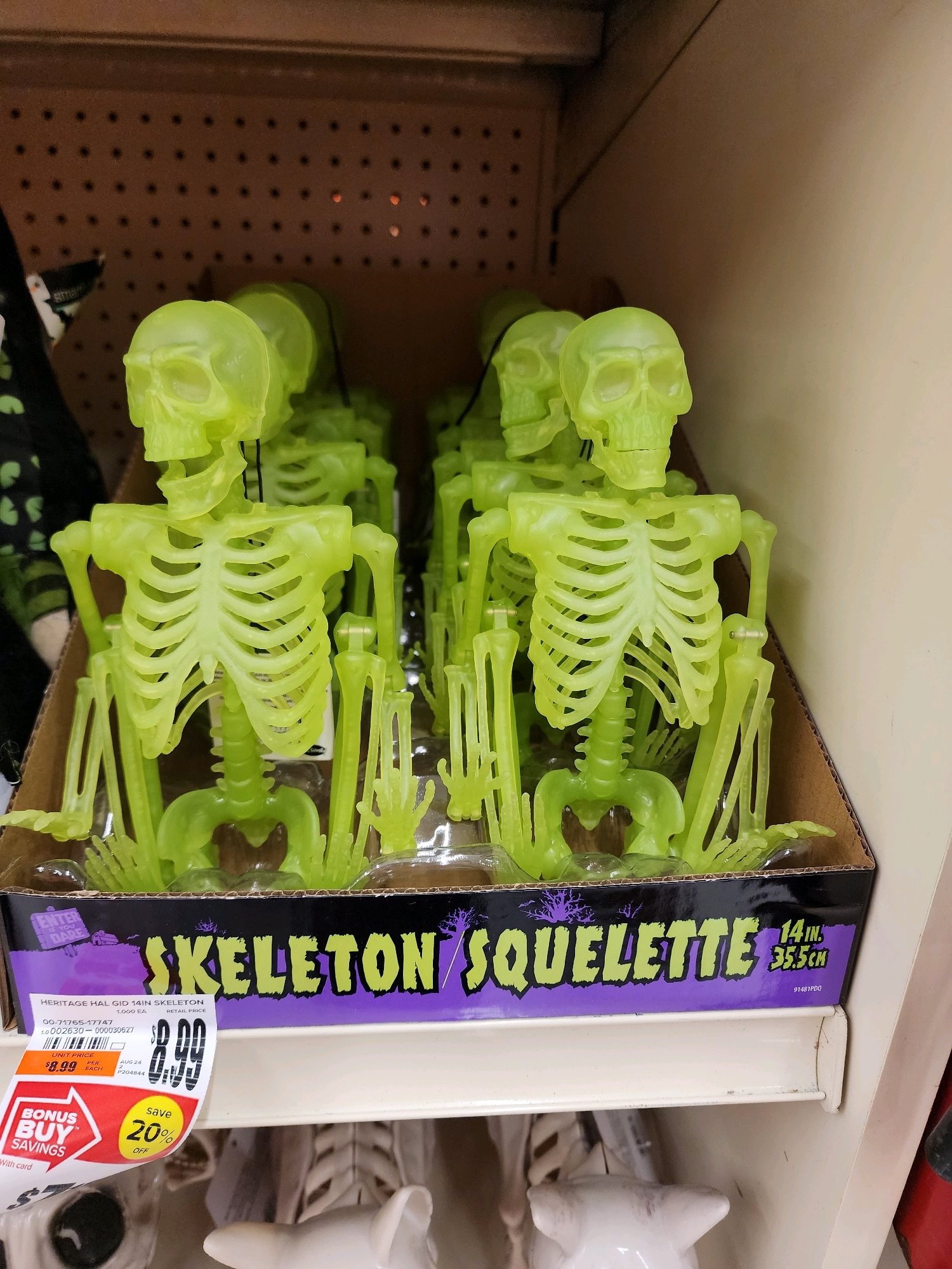 In the city of Camp Hill, United States of America, a retail store shelf displays multiple lime-green plastic skeleton figures, each approximately 14 inches tall. The skeletons are arranged in a cardboard display box with a purple and black border, featuring the text "SKELETON SQUELETTE" in a bold, spooky font. A price tag is visible on the front of the display, indicating "$8.99" for the item and a "Save 20% OFF" promotion. The figures have exaggerated skeletal features, including visible rib cages, spinal columns, and jointed limbs. The lighting in the image suggests an indoor retail environment.