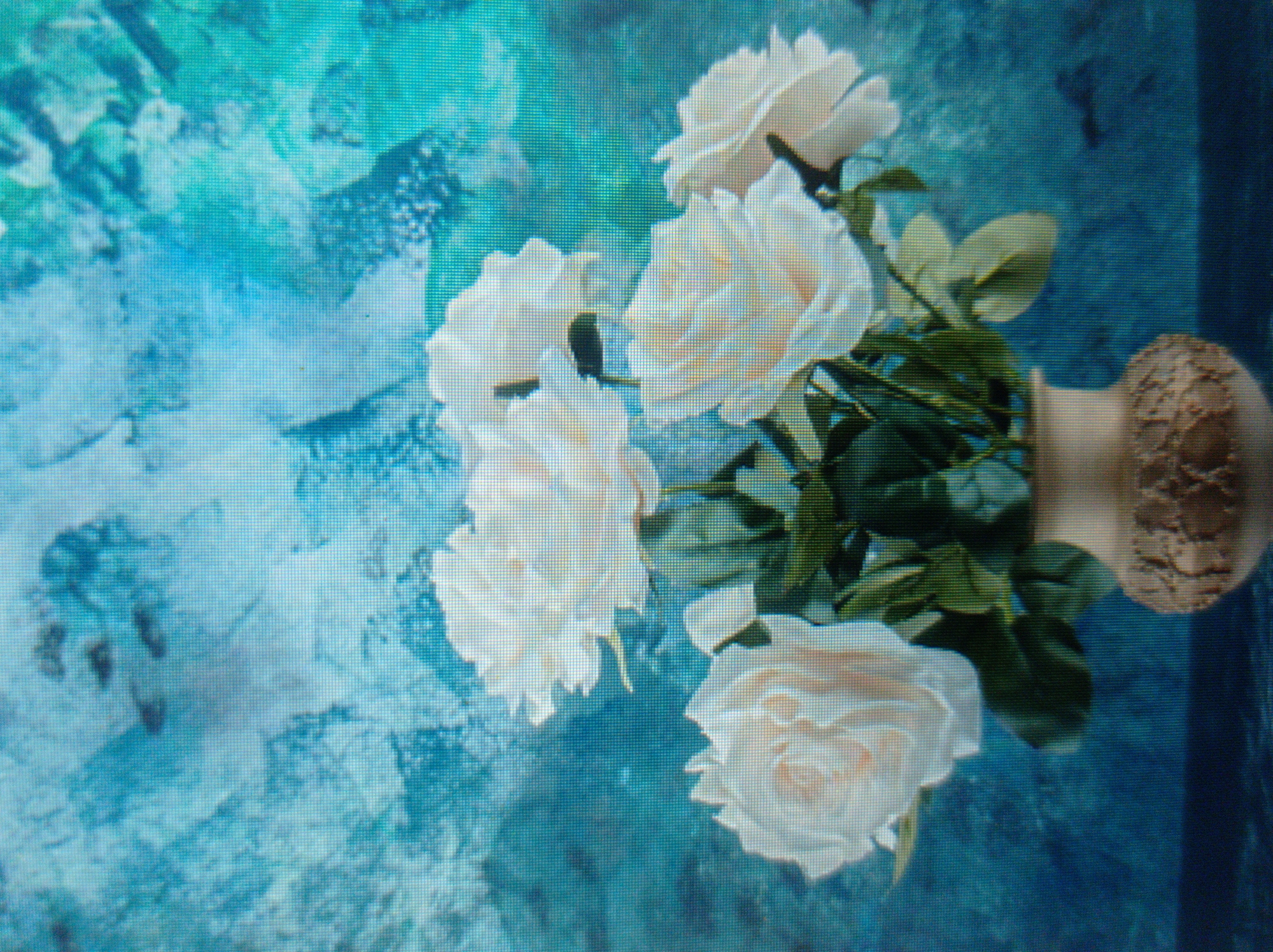 The image shows a still life arrangement of white roses in a vase against a textured blue background. The roses are in full bloom, with many petals and green leaves. The vase is light brown with an intricate pattern around its base. The background has a mottled, abstract appearance with various shades of blue and some darker shapes that could be interpreted as birds or shadows. The image is a photograph and appears to be taken indoors. There is no indication of any specific location in the city of Mukachevo, Ukraine.