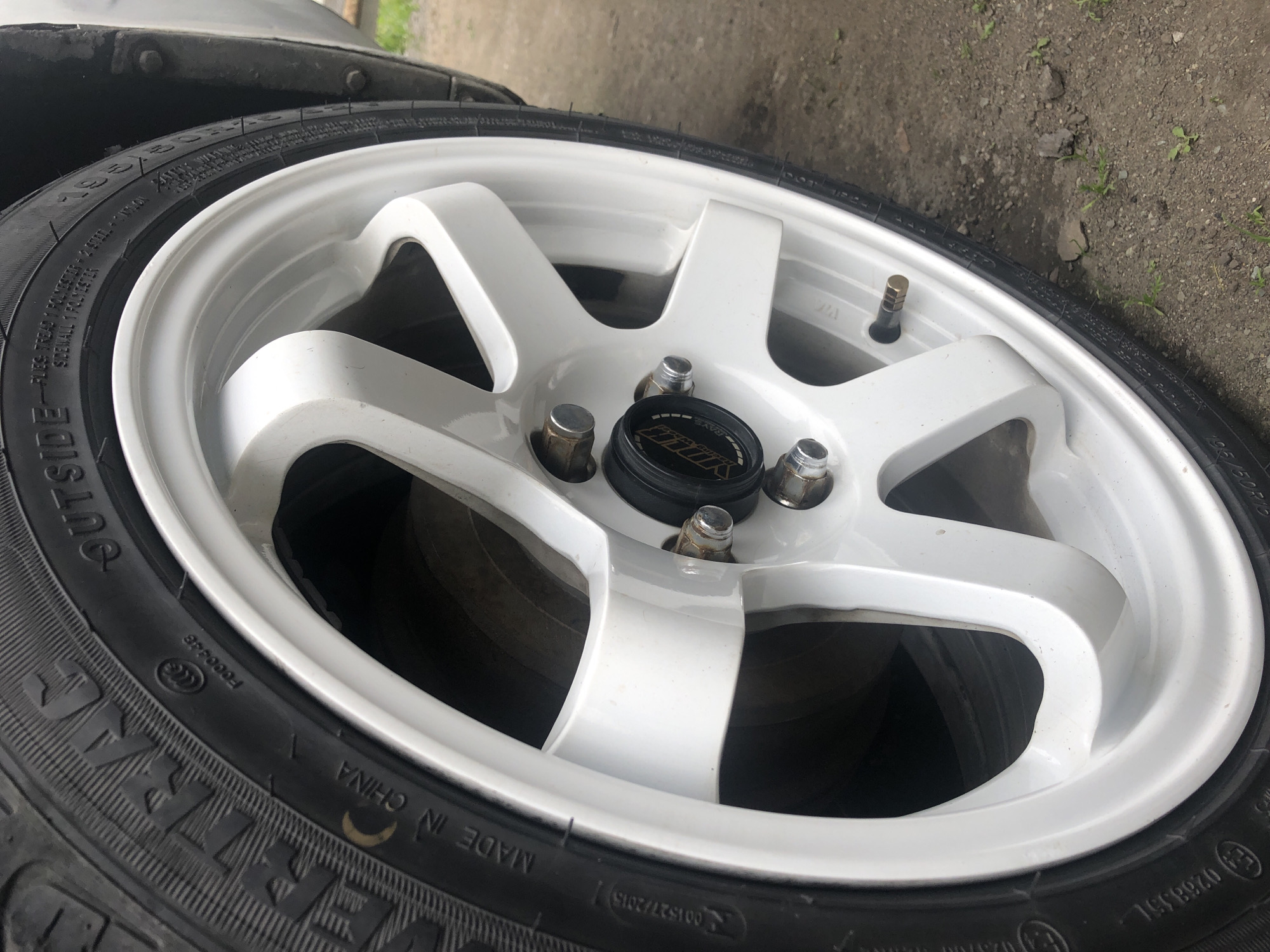 This media file shows a close-up, angled view of a white multi-spoke car wheel with a black tire. The wheel appears to be new or in good condition, with a clean, glossy finish. The tire has various markings, including "OUTSIDE," "MADE IN CHINA," and size information like "195/50R16." A black center cap with the "RAYS" logo is visible in the hub area, surrounded by lug nuts. The scene is set outdoors, with the ground appearing to be a mix of dirt and small stones. There are no people or animals visible. The lighting suggests it is daytime, possibly overcast given the lack of harsh shadows. The overall impression is a focus on the detail and appearance of the car wheel and tire.