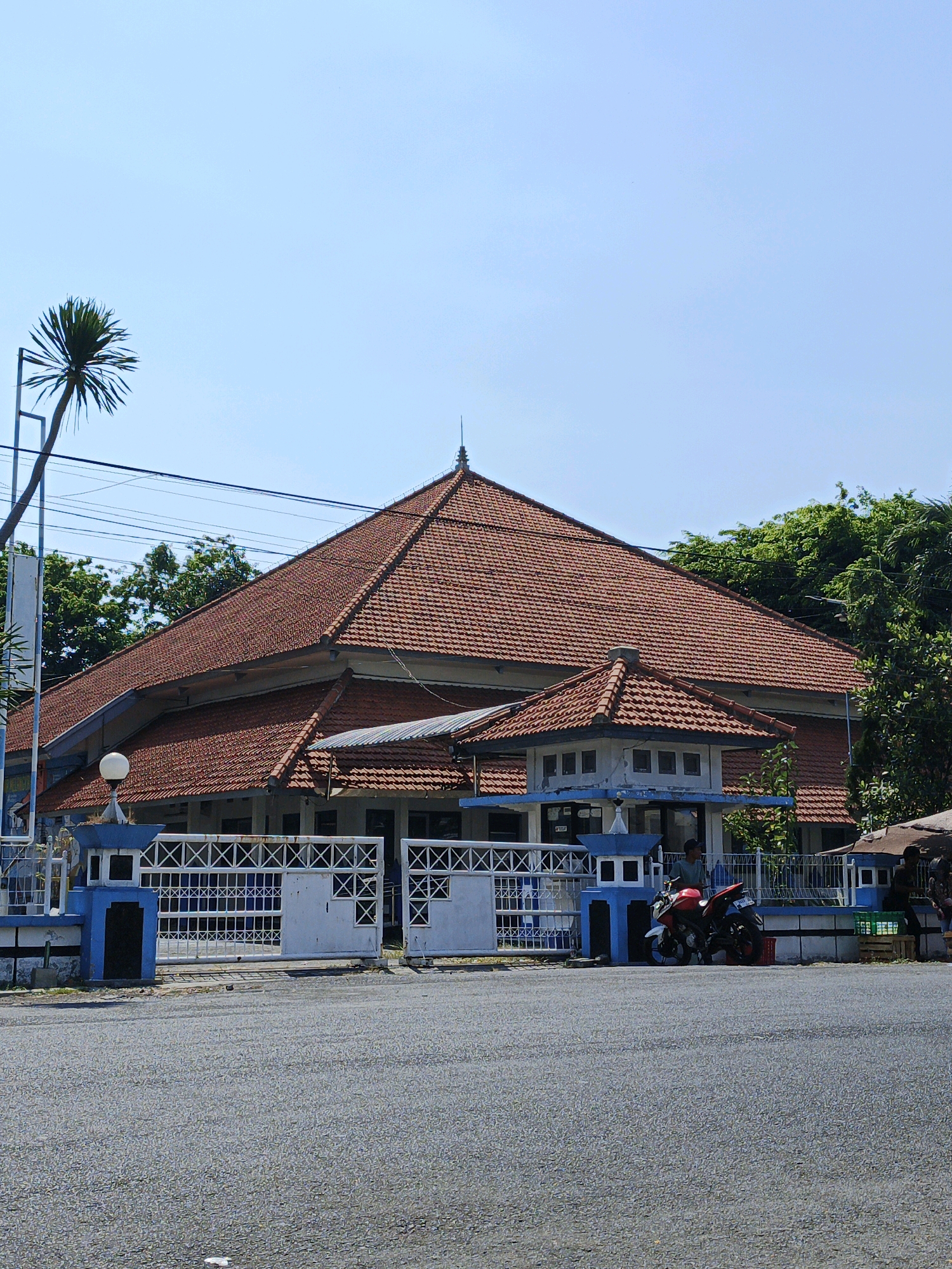 The image shows a building with a large, multi-tiered red tile roof in Kludan, Indonesia. The building is set back from a paved road and has a white fence with blue accents in front of it. There are several gates in the fence, some of which are open. A red motorcycle is parked near one of the gates. Trees and power lines are visible behind the building. The sky is clear and blue.