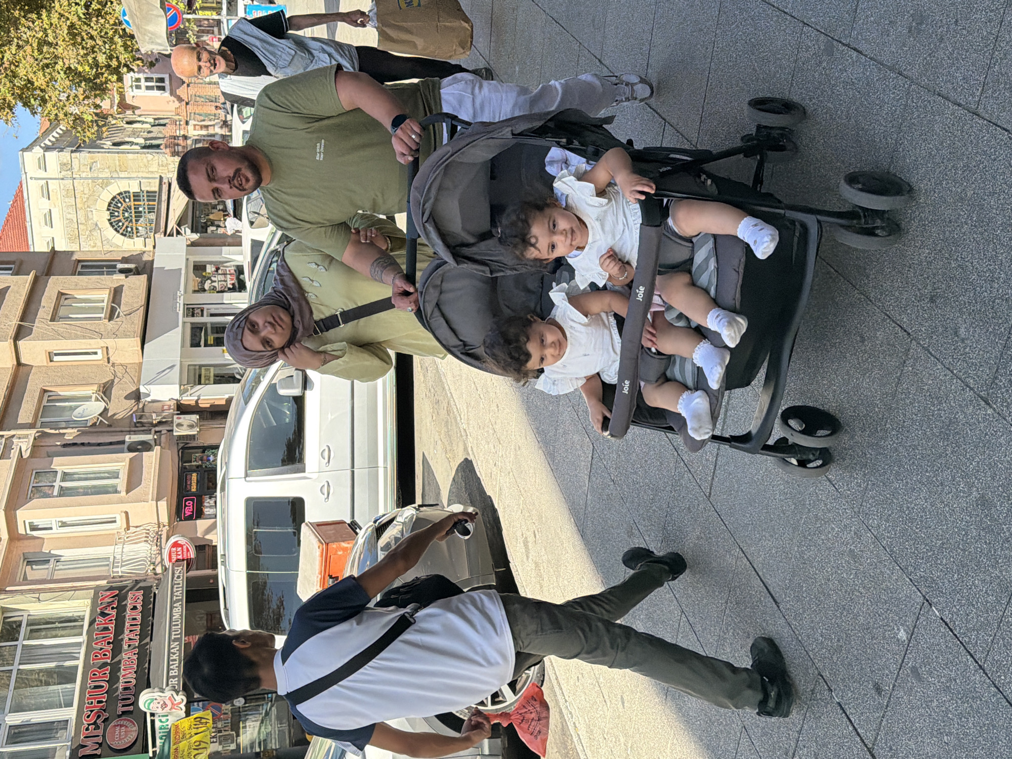 The image depicts a street scene in Kadıköy, Turkey. In the foreground, a man and woman are pushing a double stroller containing two young children. The children are wearing white outfits and white socks. The man is wearing a green t-shirt and white pants, and the woman is wearing a light green blouse and a headscarf. Behind them, an elderly man with a bald head is walking, carrying a brown paper bag. To the left, a young man in a white t-shirt and dark pants is walking away from the camera, carrying a red bag and a phone. The background shows buildings with shops, including one with signage for "Meşhur Balkan Tulumba Tatlıcısı." The street is paved with gray tiles, and there is a silver van parked on the left side of the image. The lighting suggests it is daytime, with shadows cast on the pavement.