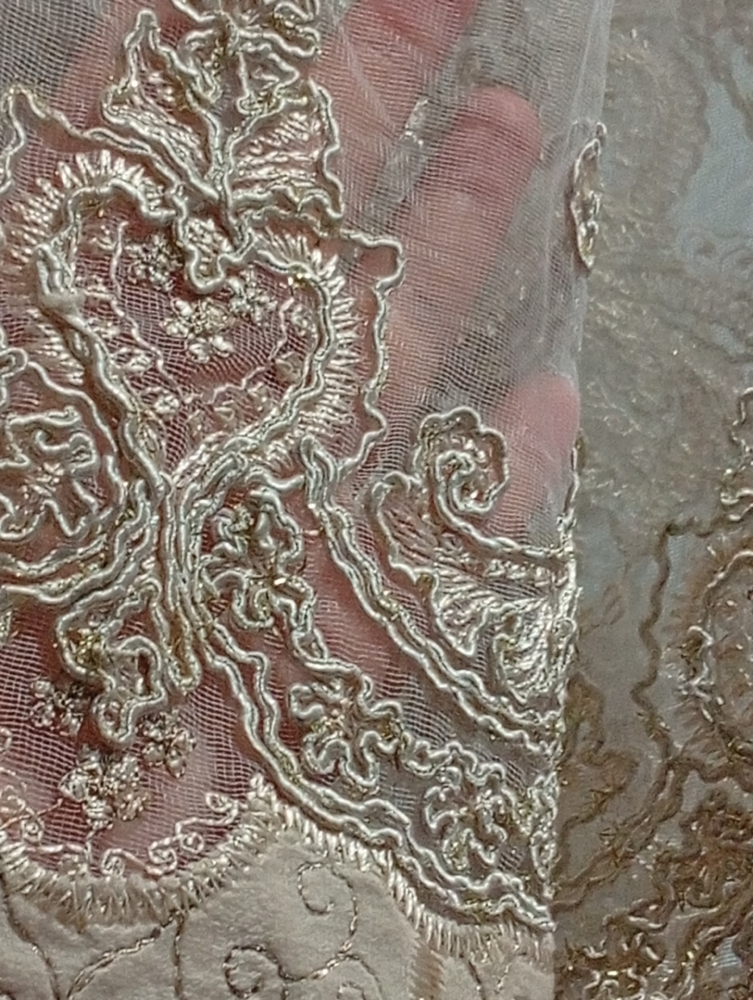 The image is a close-up, overhead shot of a piece of ornate fabric, possibly lace or embroidery, with a hand visible underneath. The fabric features intricate, raised silver thread designs on a sheer, pale pink or nude-colored mesh material. The embroidery forms swirling, floral, and leaf-like patterns. The hand underneath, showing fingers and palm, is slightly diffused by the translucent fabric, adding a sense of depth and texture to the image. The lighting appears to be soft, highlighting the metallic sheen of the embroidery threads. The focus is on the detailed craftsmanship of the fabric. There is no visible text, and the setting and time of day are not discernible beyond the fabric itself.