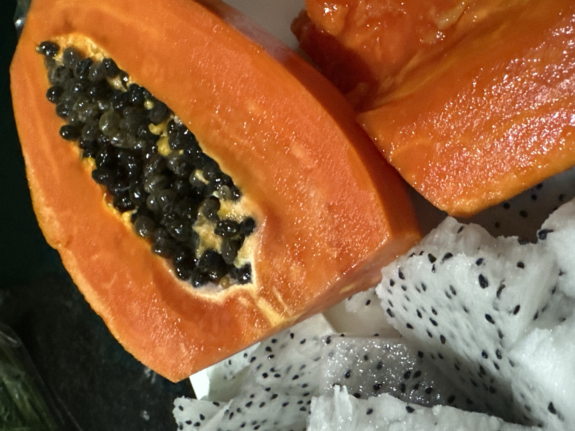The image shows a close-up of sliced fruits: papaya and dragon fruit. A section of papaya, with its orange flesh and black seeds, dominates the left side of the frame. Beside it are two slices of dragon fruit with white flesh and small black seeds. The fruits are arranged on a glossy surface, possibly a plate or tabletop. The composition is brightly lit, highlighting the textures and colors of the fruits. The city of Bac Nha Trang Ward, Vietnam is not apparent in the image.