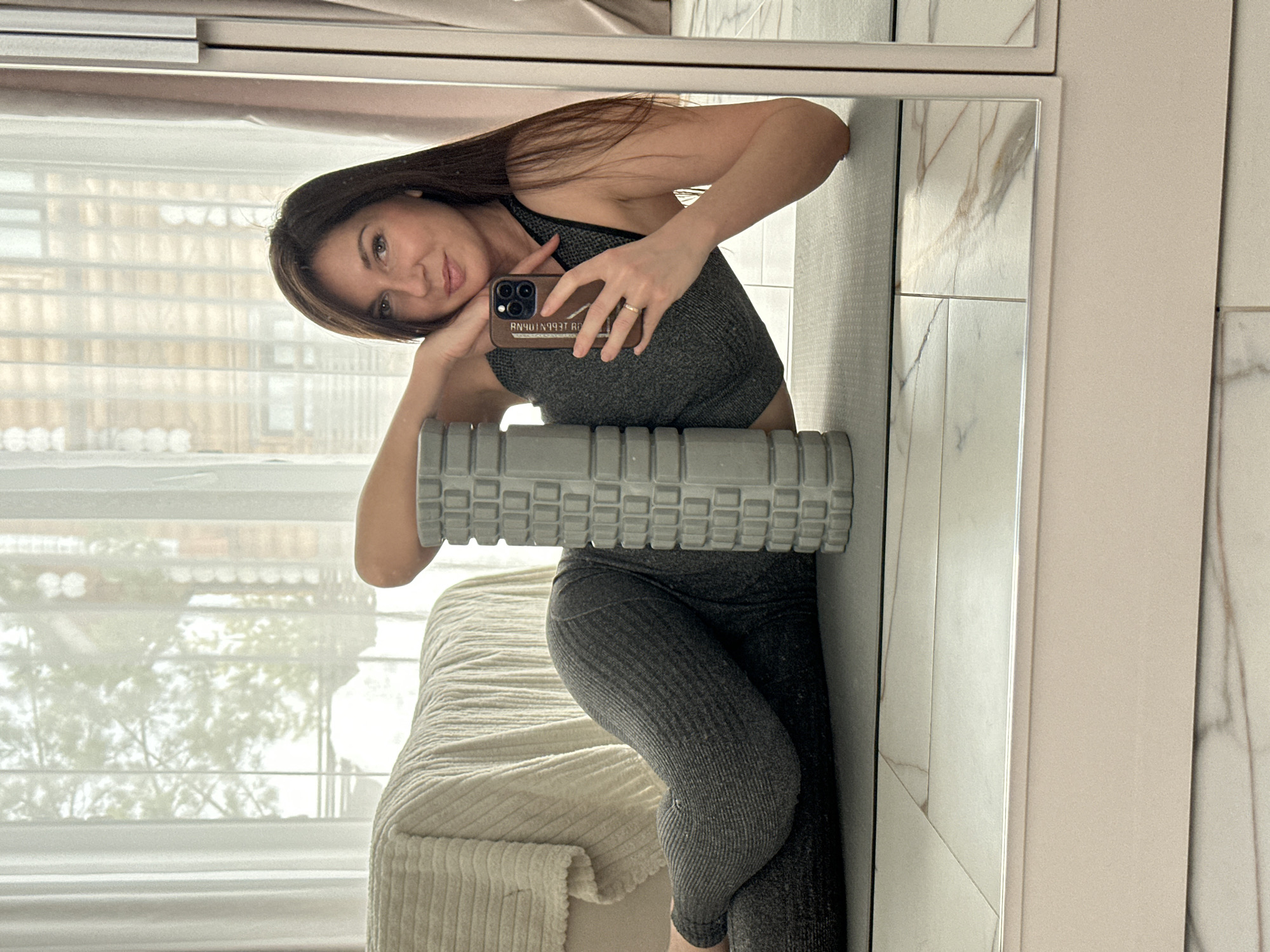 A female subject is depicted lying on her left side on a dark gray yoga mat, positioned indoors in a well-lit room. She has long, dark hair and is wearing a dark gray athletic top and matching ribbed leggings. Her left arm is bent, supporting her head, while her right hand holds a smartphone, capturing a selfie reflected in the device's screen. The smartphone has a brown case with white text, partially obscured, and she wears a ring on her left ring finger. A gray foam roller with a textured, grid-like surface is positioned vertically behind her back, providing support as she leans against it. To the left of the subject, a light-colored, textured throw blanket covers furniture, likely a bed or sofa. Behind this furniture, a large window is covered by sheer white curtains, allowing diffused natural light into the room. Blurred green foliage and some building structures are faintly visible through the curtains. The floor is covered with light-toned tiles featuring subtle brown and white veining. A large mirror on the right side of the frame reflects the floor and parts of the room. The specific city location of Chaikovsky, Russia, cannot be confirmed from the visual evidence.