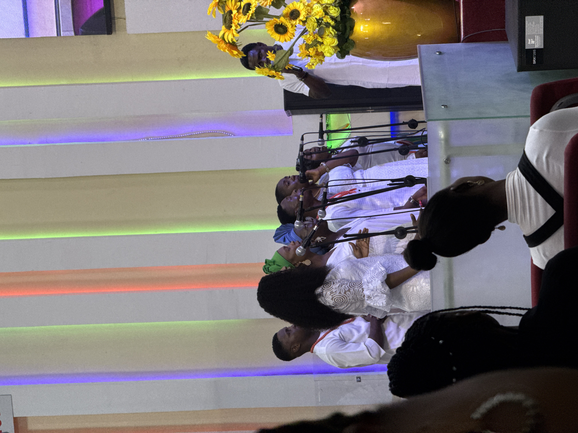 In a brightly lit interior, possibly a church or auditorium in Lugbe, Nigeria, a choir of approximately six individuals stands behind microphones. They are dressed in white attire and appear to be singing. A man is standing behind them, tending to a large arrangement of sunflowers in a vase. The backdrop features horizontal bands of colored lights: orange, green, blue, and yellow. A section of the audience is visible in the foreground.