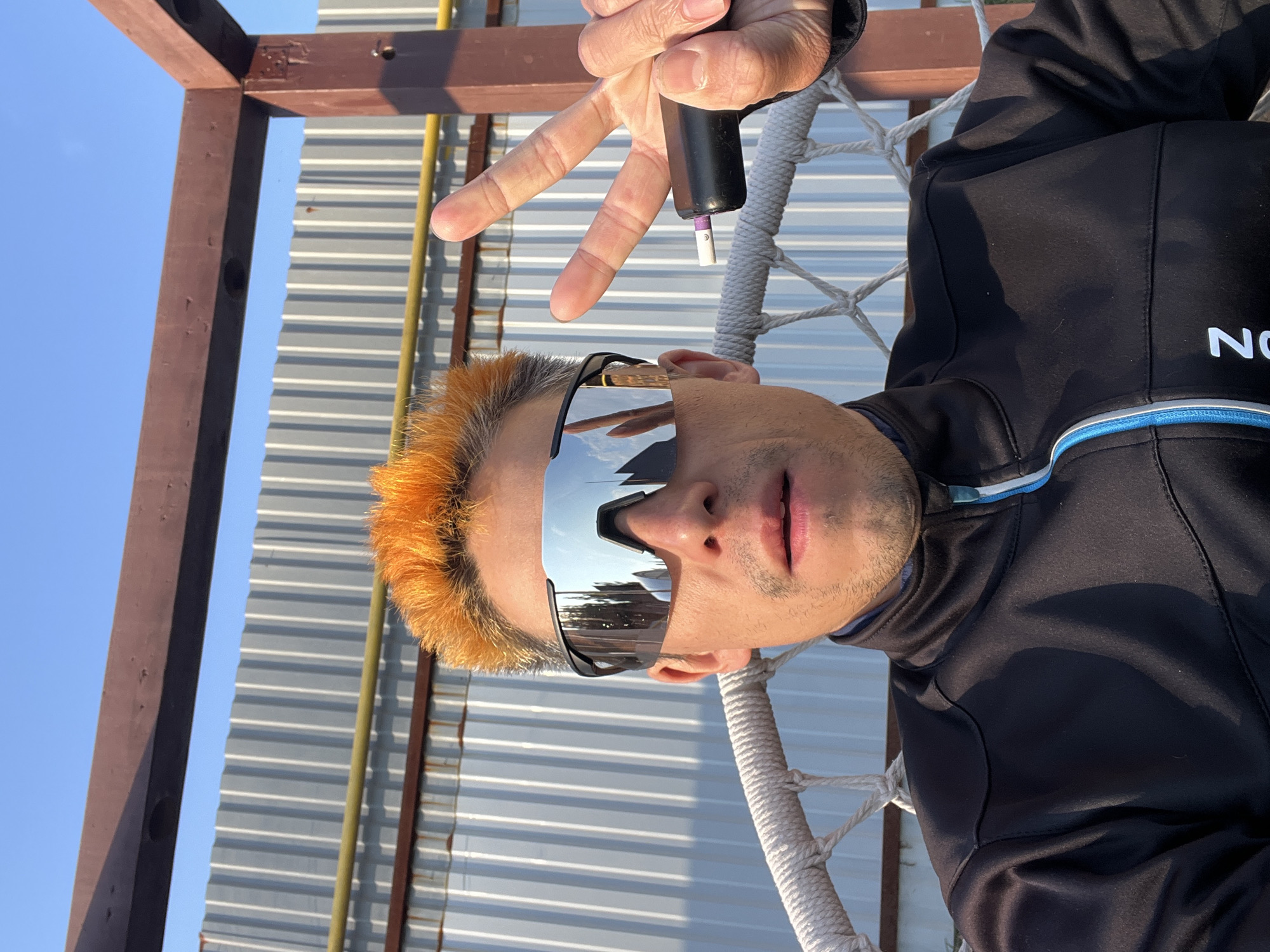 A man sits in a white macrame chair, with a corrugated metal structure and a brown wooden frame behind him. He has orange hair, mirrored sunglasses, and is holding an electronic cigarette in his hand, making a hand gesture with his other hand. He is wearing a black jacket with a white logo. The location is in the city of Chaikovsky, Russia.