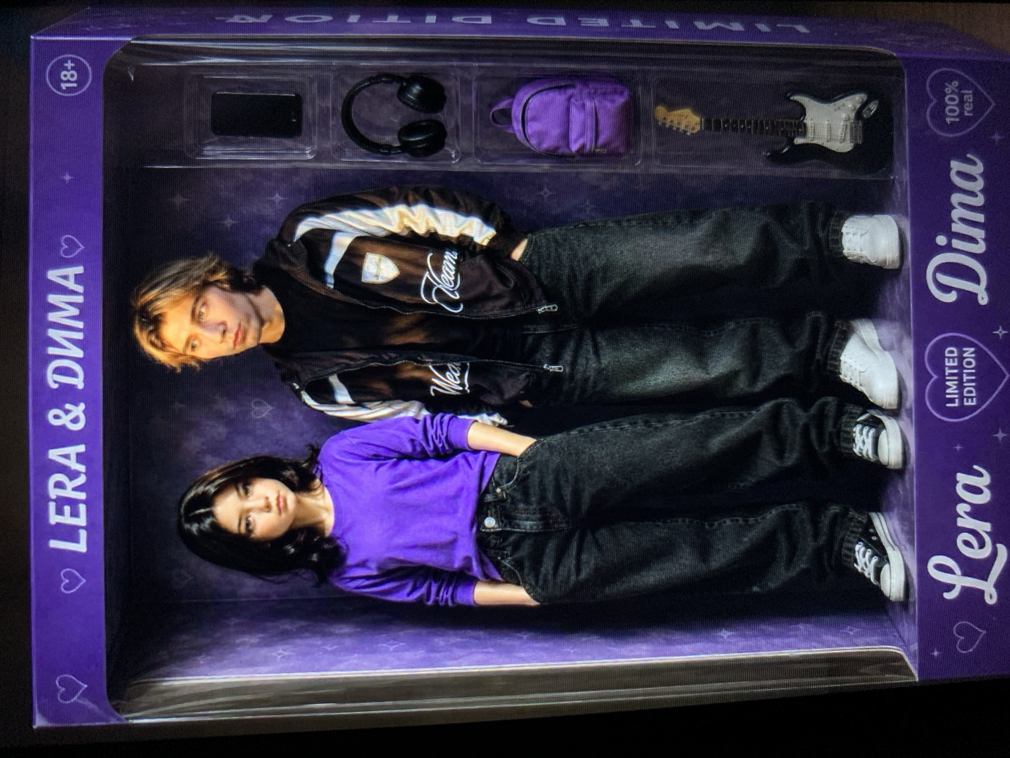 The image shows a doll box with two figures, Lera and Dima, inside. The box is purple and has text in white, including "LERA & DIMA" at the top and "Lera" and "Dima" at the bottom. There are accessories in the box, such as a phone, headphones, a backpack, and a guitar. The figures are dressed in casual clothing. Lera is wearing a purple long-sleeved shirt and black jeans, while Dima is wearing a black jacket with white stripes and black jeans. The overall presentation suggests a limited edition collectible item. The image itself is a photograph, taken indoors with artificial lighting. There is no indication of Lymanka, Ukraine, in the image or its content.
