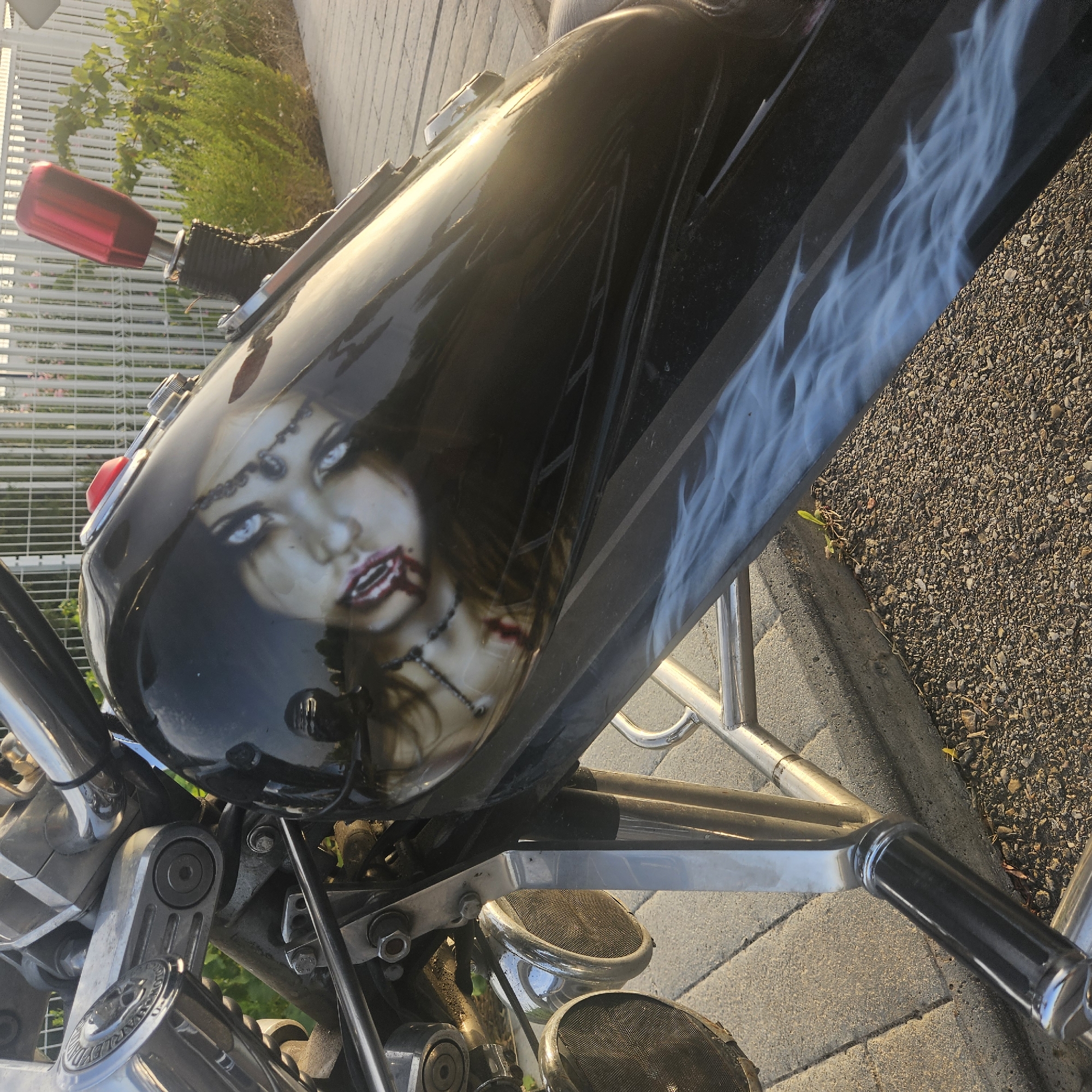 The image shows a close-up of a custom motorcycle's fuel tank, which features an airbrushed design of a woman's face with striking blue eyes and blood dripping from her mouth. The motorcycle is parked outdoors on a paved surface, possibly a sidewalk or driveway, with a white metal fence and green foliage visible in the background. The sun is casting warm light on the scene, reflecting off the motorcycle's glossy black paint. The motorcycle appears to be a cruiser style, with chrome accents and custom detailing. The surrounding environment suggests a sunny day in a residential or urban area. There are no people present in the immediate vicinity.