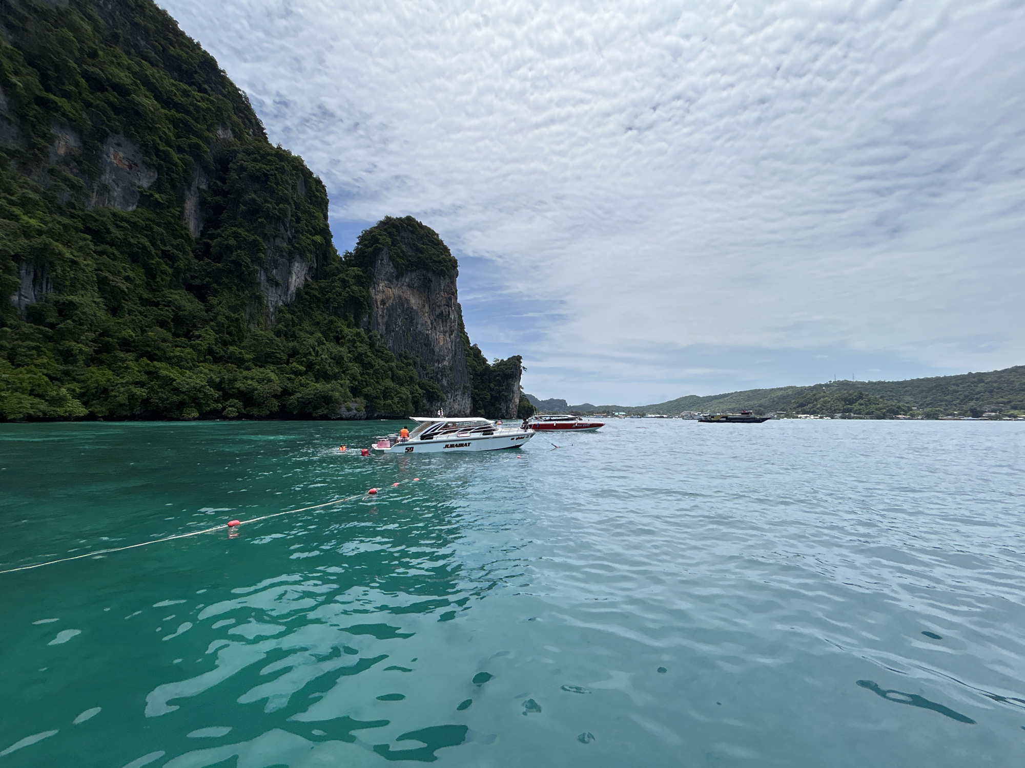In Ao Nang, Thailand, a wide-angle shot captures a serene seascape under a cloudy sky. The turquoise water is calm, with gentle ripples disturbing its surface. A white speed boat, bearing the name "JURAIRAT" and the number "59," is docked in the foreground, its white canopy providing shade. A rope with red buoys stretches across the water, marking a boundary or perhaps a swimming area. In the background, a red speed boat is visible, as well as a ferryboat further in the distance. Towering limestone cliffs, covered in lush green vegetation, rise majestically on the left side of the frame, their imposing presence dominating the landscape. The horizon is dotted with more islands and a hint of land with structures on the right. The overall scene evokes a sense of tranquility and natural beauty.
