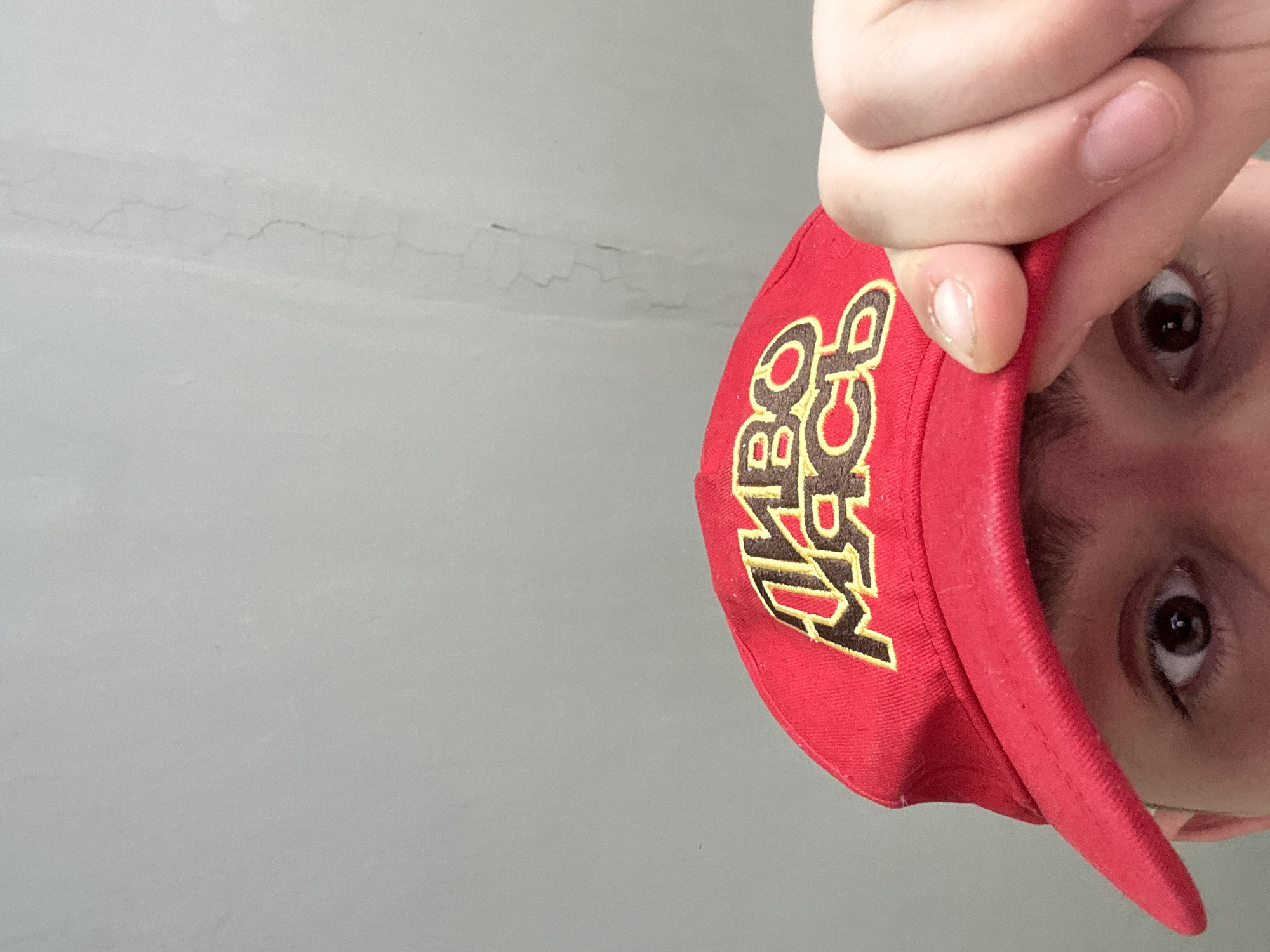 The image shows a person wearing a red baseball cap with yellow and brown embroidery, likely taken from a low angle. The person's eyes and part of their face are visible. The background appears to be a light grey wall. The location is not specified in the image.