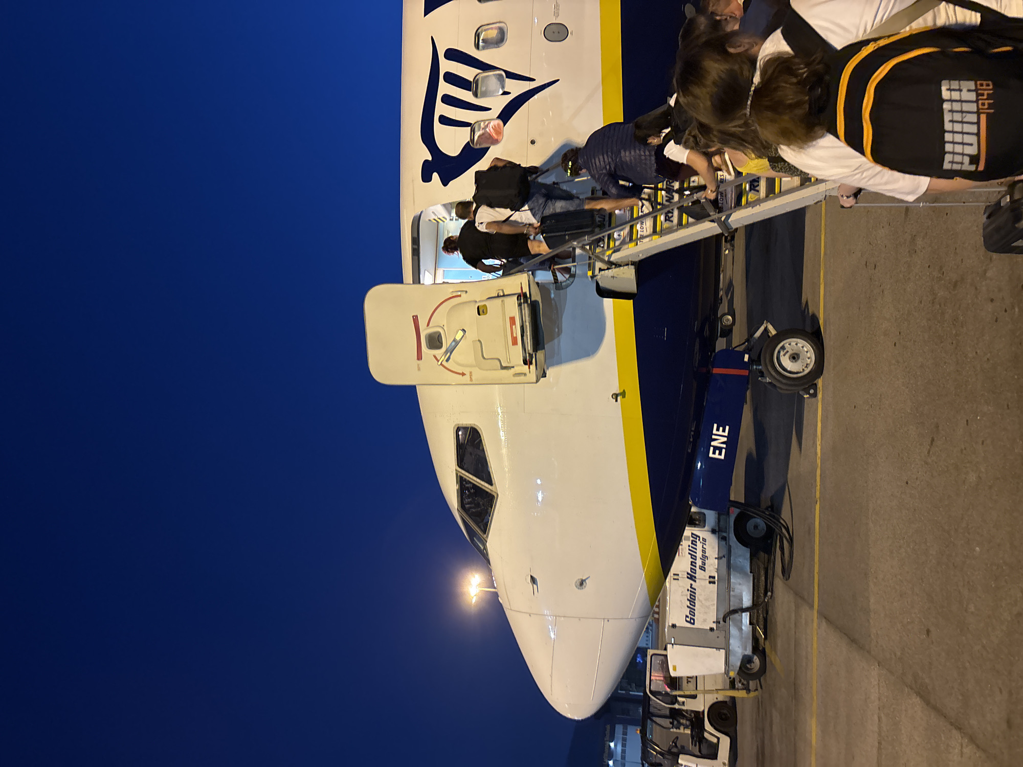 This image depicts a nighttime scene at an airport in Sofia, Bulgaria. The focus is on the front section of a Ryanair airplane, with its iconic blue and yellow livery visible. A set of mobile stairs is attached to the aircraft's open door, and passengers are boarding.
In the foreground, several passengers are seen from behind as they ascend the stairs. One person is wearing a black backpack with an orange trim and the "Puma 1948" logo. Another passenger further up the stairs carries a black rolling suitcase.
On the tarmac, a ground support equipment unit is positioned near the aircraft, bearing the text "Goldair Handling Bulgaria." A small airport tug or cart is also visible to the left. The sky is a deep, dark blue, suggesting late evening or early night. The airport environment is characterized by artificial lighting, including a bright spotlight illuminating the cockpit area of the airplane. The scene conveys the activity of boarding an aircraft, likely for a flight departing from Sofia.