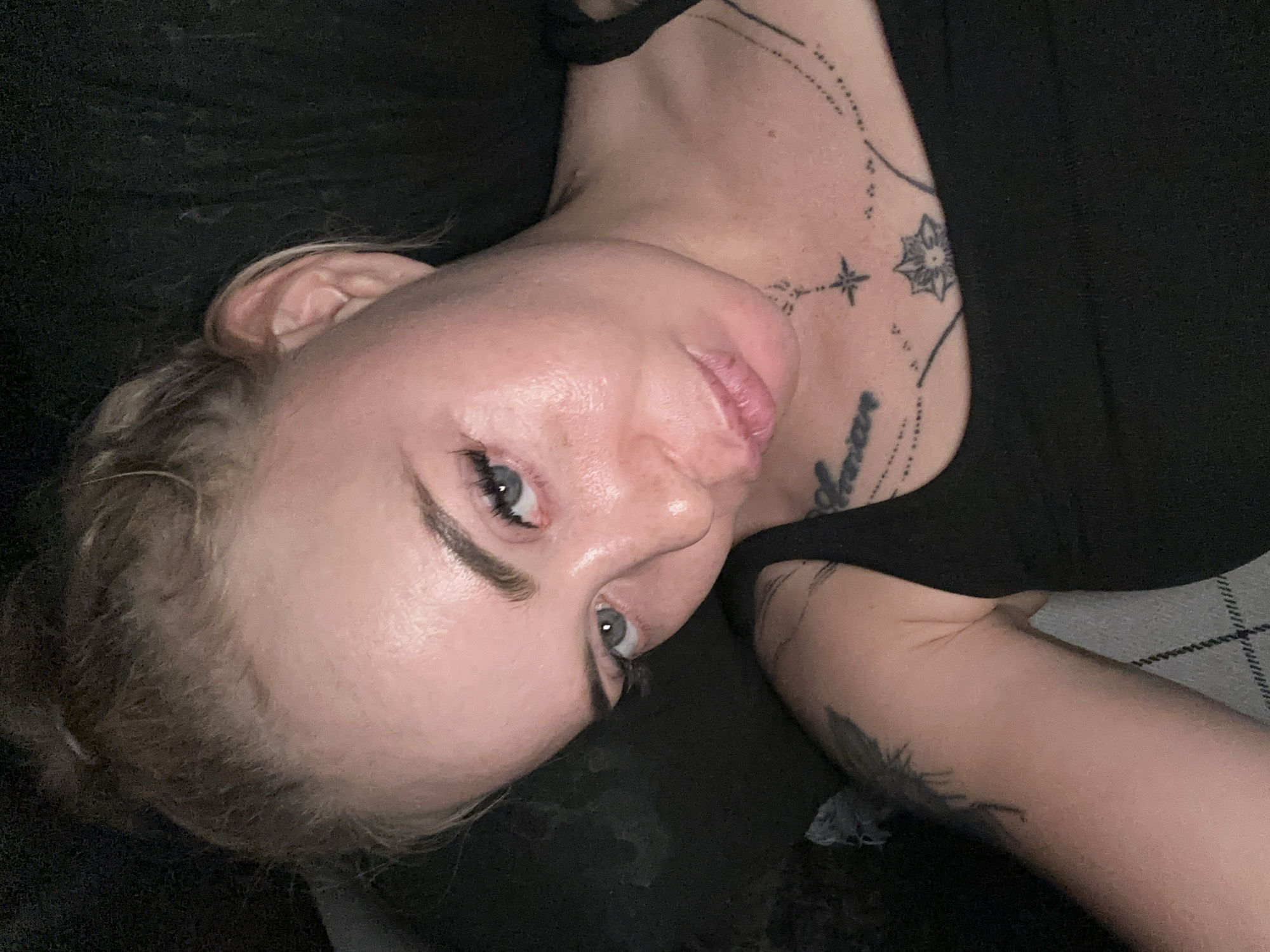 A woman with her hair pulled up in a messy bun lies on a dark surface, looking towards the camera. She has light blue eyes, fair skin, and is wearing a black tank top. Tattoos are visible on her chest and left arm. The lighting is dim, and the background is dark and indistinct.