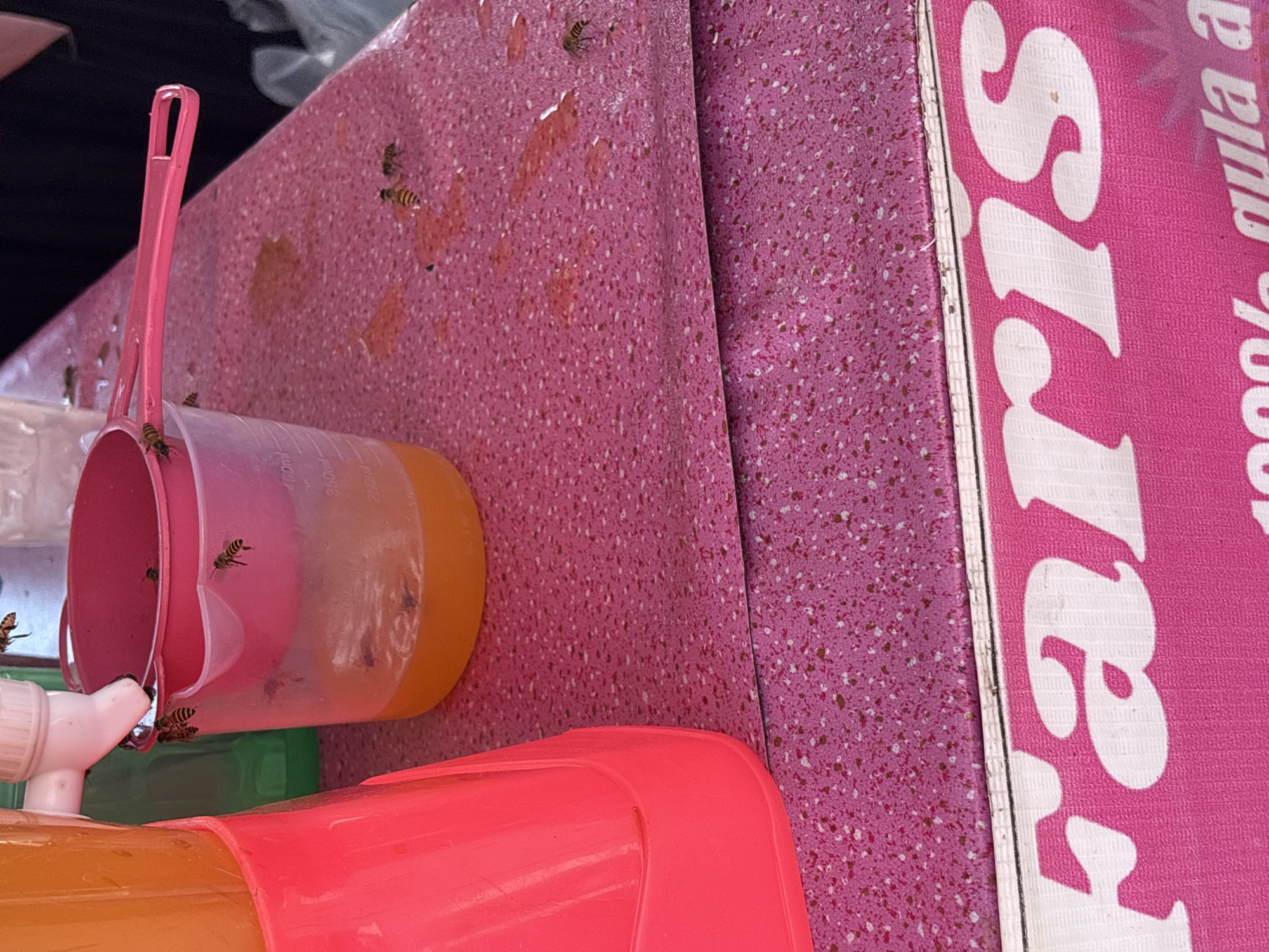 The image depicts a close-up, overhead view of a pink speckled countertop, likely outdoors at a street vendor stall in Lhokseumawe, Indonesia, given the presence of the sign with Indonesian text. A bright pink dispenser on the left holds an orange liquid, possibly juice. To the right of the dispenser is a pink plastic measuring cup containing more orange liquid and a pink plastic scoop. Several bees are attracted to the liquid, with some on the rim of the cup, the scoop, and the countertop. A green bottle is visible behind the measuring cup. The lower portion of the image shows a pink banner with white text, which appears to be a brand name or product advertisement. The overall scene suggests a refreshing drink being sold, unfortunately attracting unwanted insect visitors.
