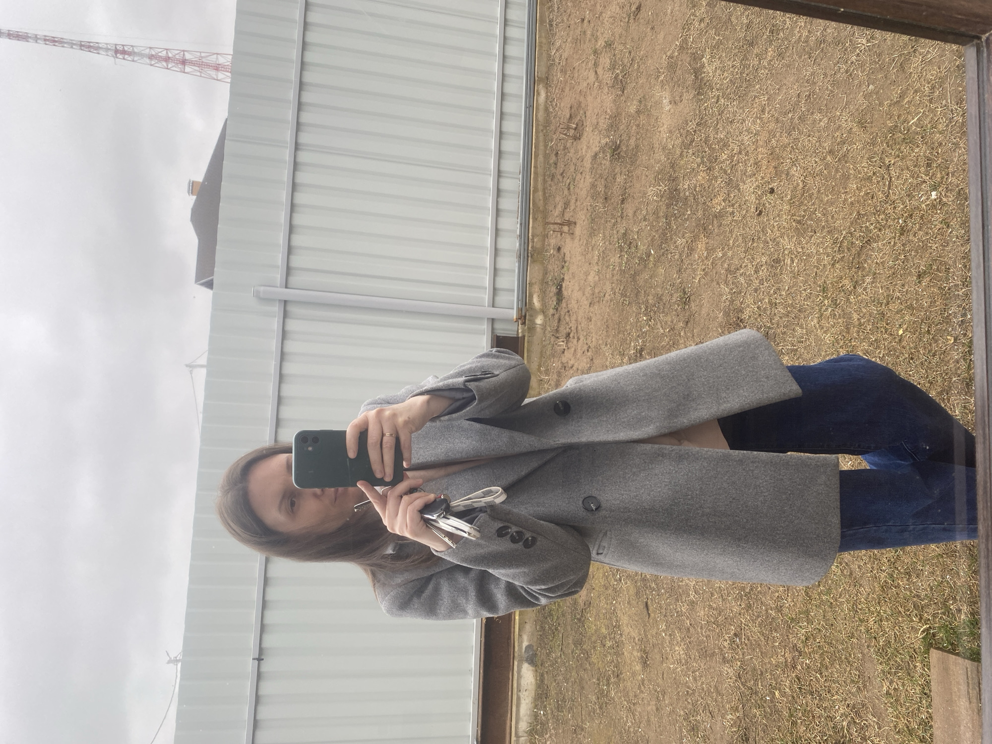A woman is taking a selfie in a mirror, holding a smartphone with a green case in her right hand and a set of keys in her left. She is wearing a gray overcoat and blue jeans. The reflection shows an outdoor scene with a gray metal fence running horizontally across the top half of the frame. Above the fence, a red and white lattice tower is visible against a cloudy sky. The ground is dry and covered with brown grass. A building with a dark-colored roof is partially visible behind the fence. The mirror frame is visible on the right side of the image. The scene is in Chaikovsky, Russia.
