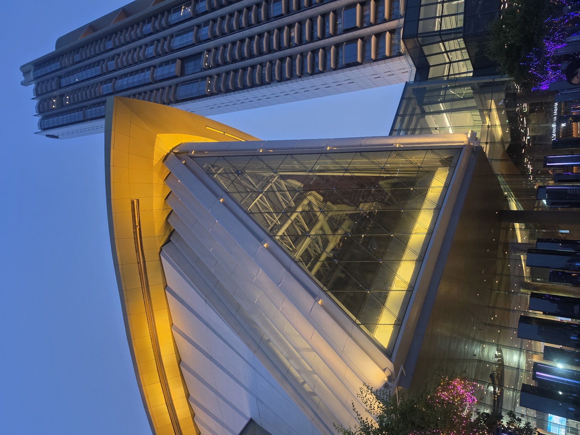 This image depicts the architectural details of a modern building complex in Bangkok, Thailand, taken during twilight. The scene focuses on three distinct structures. At the top, a multi-story building with horizontally-arranged architectural elements dominates the skyline. Below it, a bright yellow, curved structure connects to a transparent, triangular glass structure with a visible support network inside. To the right, is a flat, dark glass building, reflecting nearby lights, and including some sparse foliage, and a few hints of activity on the sidewalk. The sky is a gradient of blue hues. There are no people visible in the main focus area, though a face is visible on the right edge of the photo.
