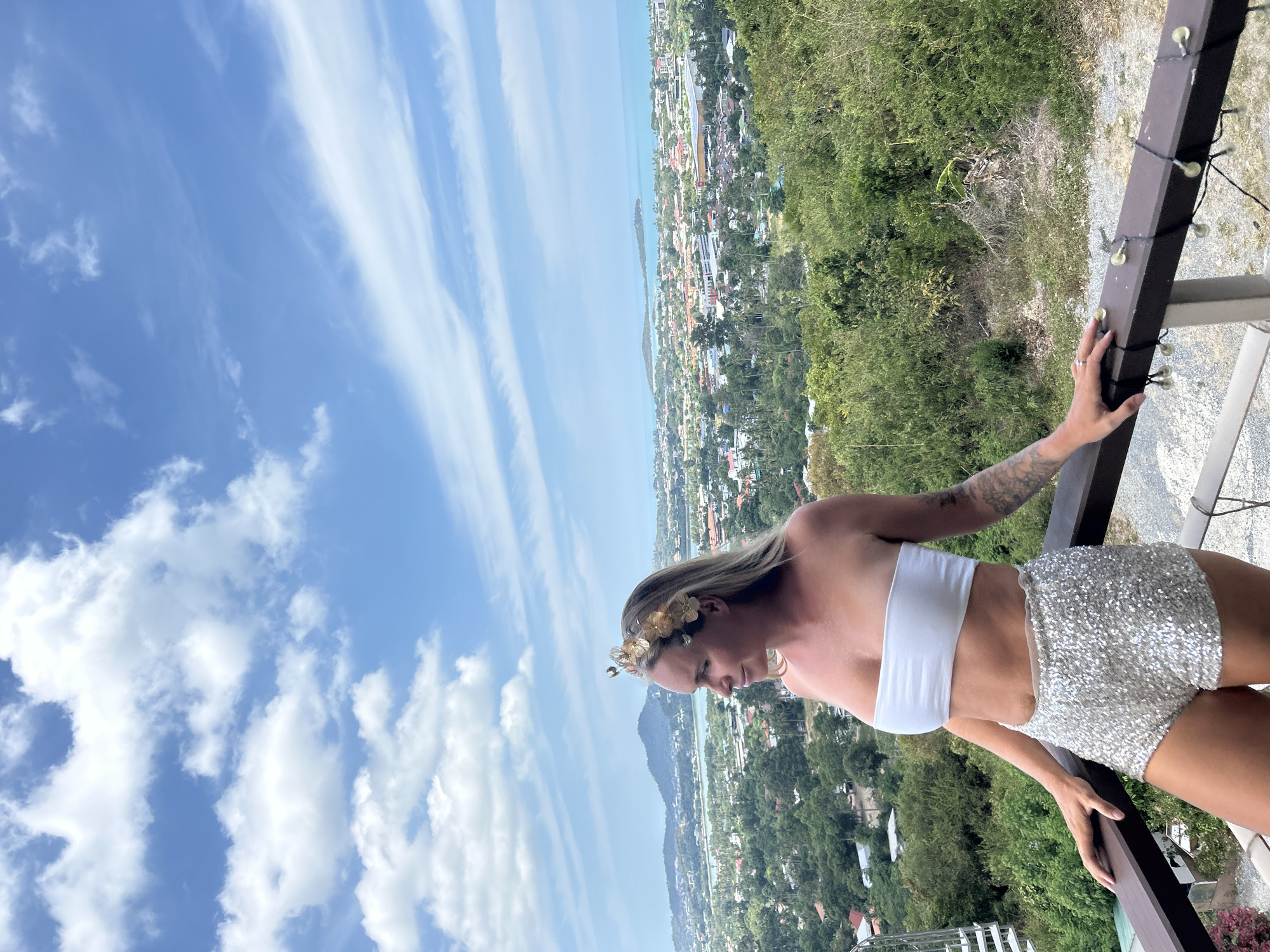 An outdoor, medium-shot, eye-level, waist-up photograph shows a blonde woman with a gold flower crown on her head and a white bandeau top, standing on a balcony overlooking a city and the ocean. The woman is wearing sparkly, silver shorts. She is facing right and looking out at the horizon. The balcony railing in front of her is dark brown and adorned with string lights. In the background, a lush green landscape with buildings and trees leads to a body of blue water, with mountains and the sky visible beyond. The sky is bright blue with scattered white clouds. The location is the Ko Samui District in Thailand, as indicated by the tropical vegetation and architecture.