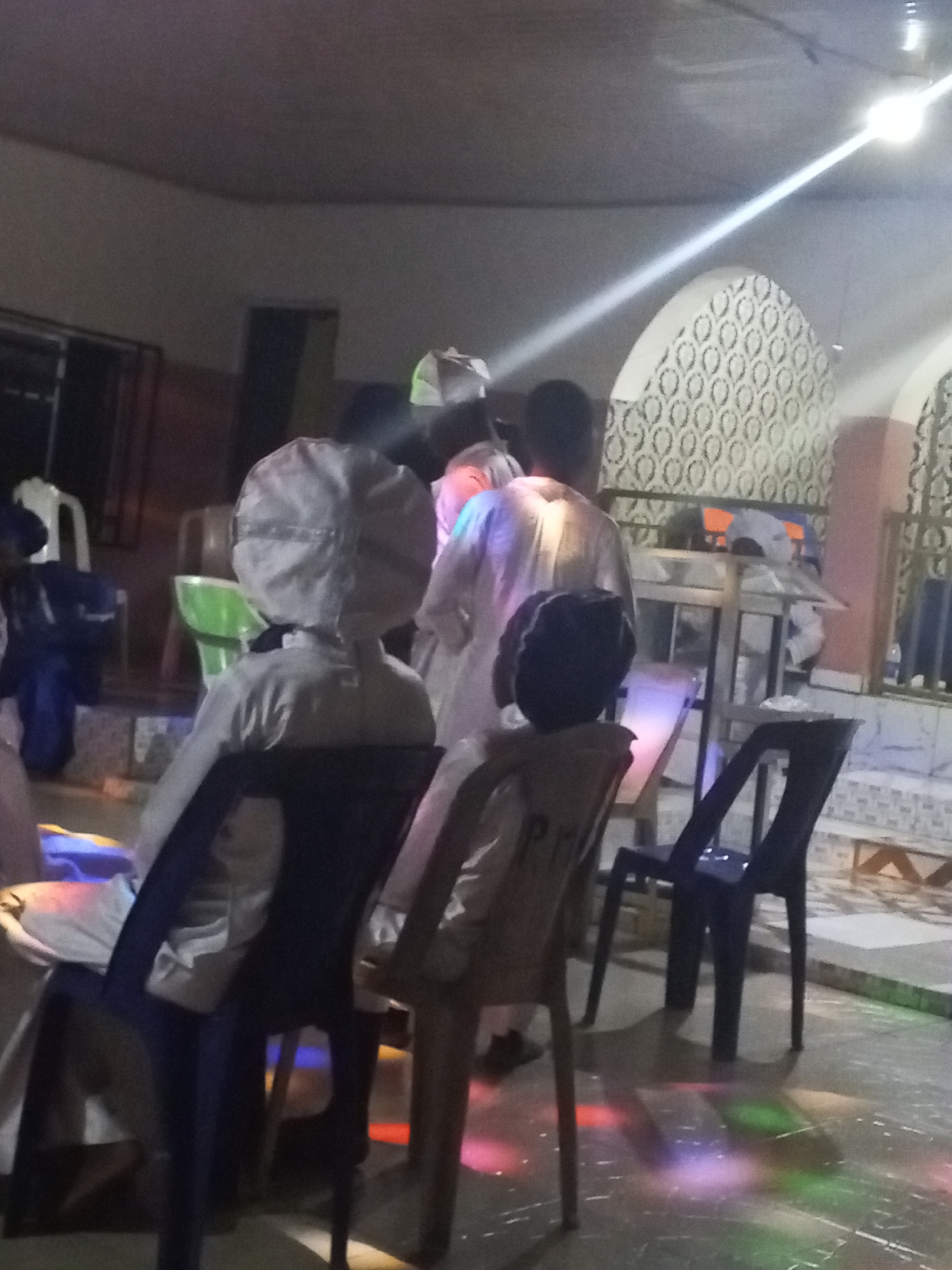 In a dimly lit room, possibly in Lokoja, Nigeria, several people are observed from behind. They are dressed in what appears to be white robes and head coverings. Some are seated on plastic chairs, while others are standing. The room features arched windows with decorative patterns, and a metallic structure is positioned near the back. The floor is marked by colorful light patterns.