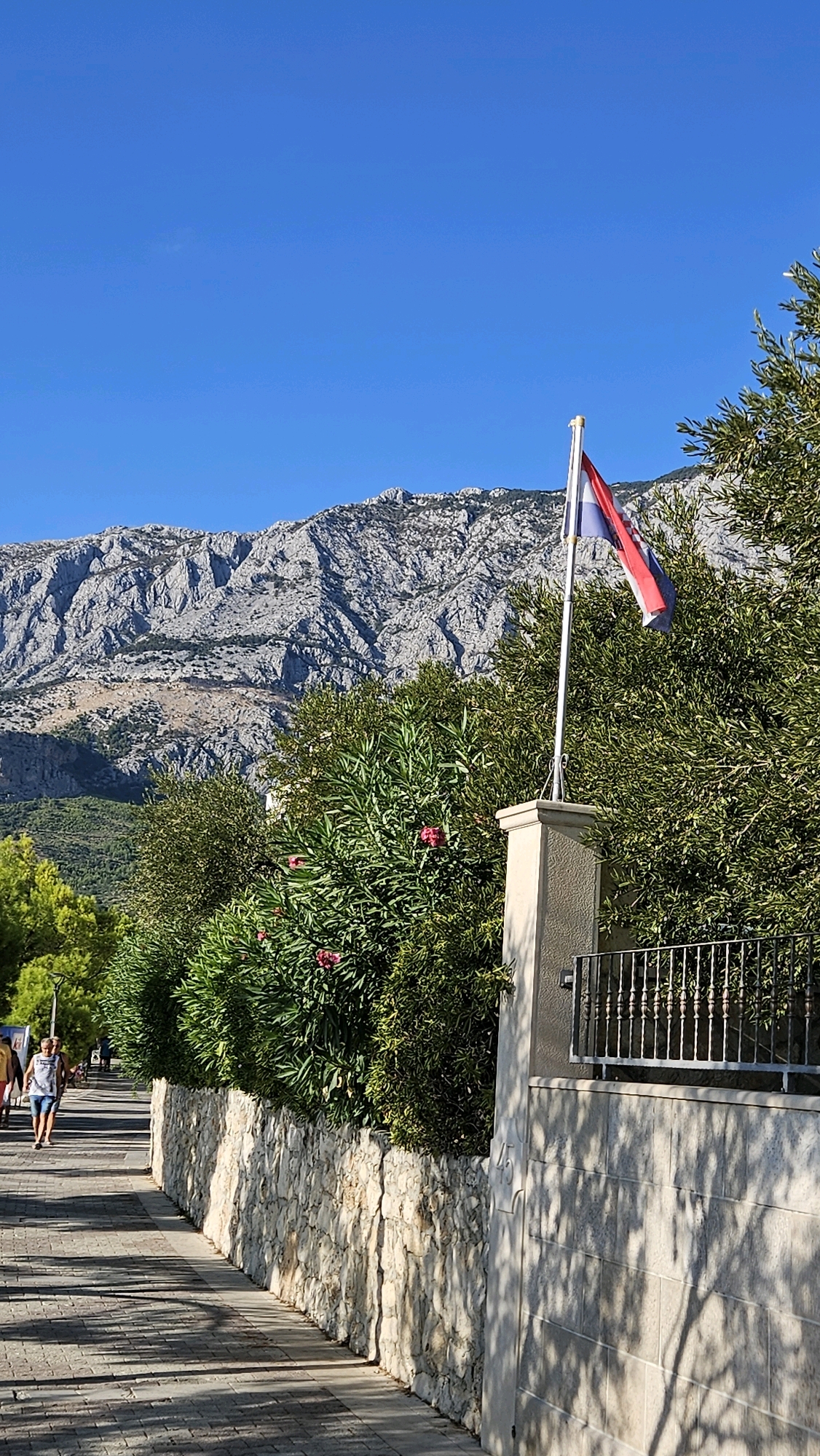 In Općina Tučepi, Croatia, a paved walkway runs parallel to a textured stone wall topped with dense green foliage. A flag, red, white, and blue, flies from a pole beside a stone pillar on the right side of the frame. Behind the foliage and wall, a rugged, rocky mountain range dominates the background under a clear blue sky. Several people are visible on the walkway in the distance, walking in the same direction, with one individual in the foreground wearing a white tank top and blue shorts. Sunlight casts dappled shadows from the surrounding trees onto the walkway and the stone wall.