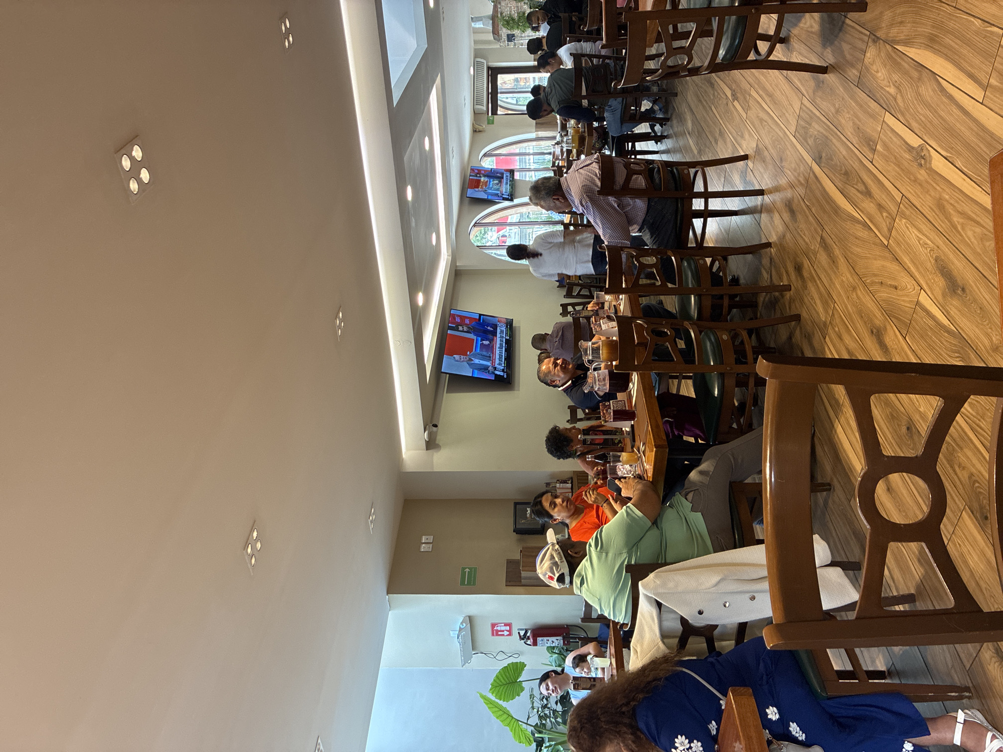 A restaurant scene in Cuautitlán Izcalli, Mexico, shows a long, narrow dining area with rows of wooden tables and chairs occupied by patrons. The wooden floor complements the brown furniture. Several television screens are mounted on the walls, displaying content. People are seated at the tables, appearing to be engaged in conversations or dining. Interior lighting fixtures are visible on the ceiling. A plant is at the near end of the tables. A fire extinguisher is mounted on the wall.