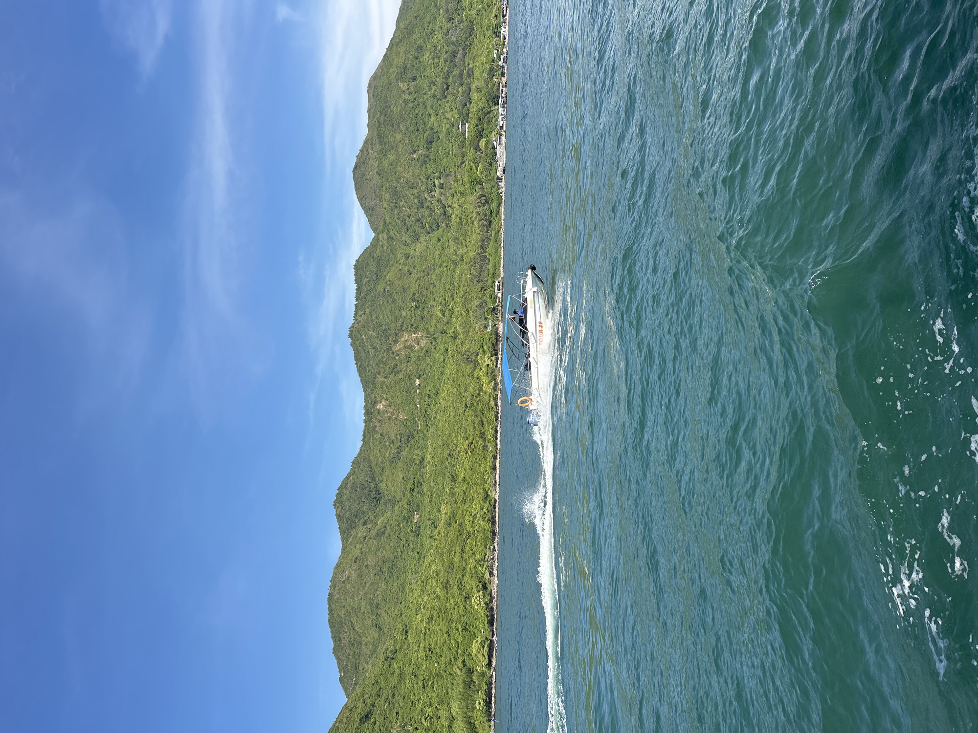 A white speedboat with a blue canopy is moving through choppy turquoise water, leaving a white wake behind it. The boat has a few people visible inside and appears to be on a tour, with "TOUR 29" written on its side. In the background, lush green, tree-covered mountains rise steeply from the coastline. The sky is a clear, bright blue with a few wispy clouds. The scene is set on the coast of Nha Trang, Vietnam, likely near a bay or island.