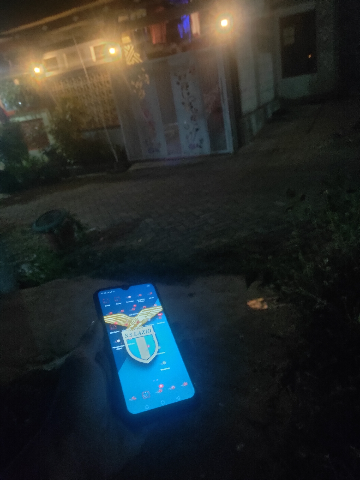 The image shows a close-up of a smartphone held in a person's hand at night, in what appears to be an outdoor residential area. The smartphone screen displays a blue background with the S.S. Lazio crest as a prominent icon. The crest features an eagle with outstretched wings above a shield divided into green, white, and blue sections, with "S.S. LAZIO" written below it. Several other app icons are visible on the screen. The background of the image is dark and out of focus, but suggests a paved pathway or ground leading towards a building with lit windows and a fence or gate. The overall lighting suggests it is evening or night, with artificial lights illuminating parts of the scene. The location is stated to be Antirogo, Indonesia.
