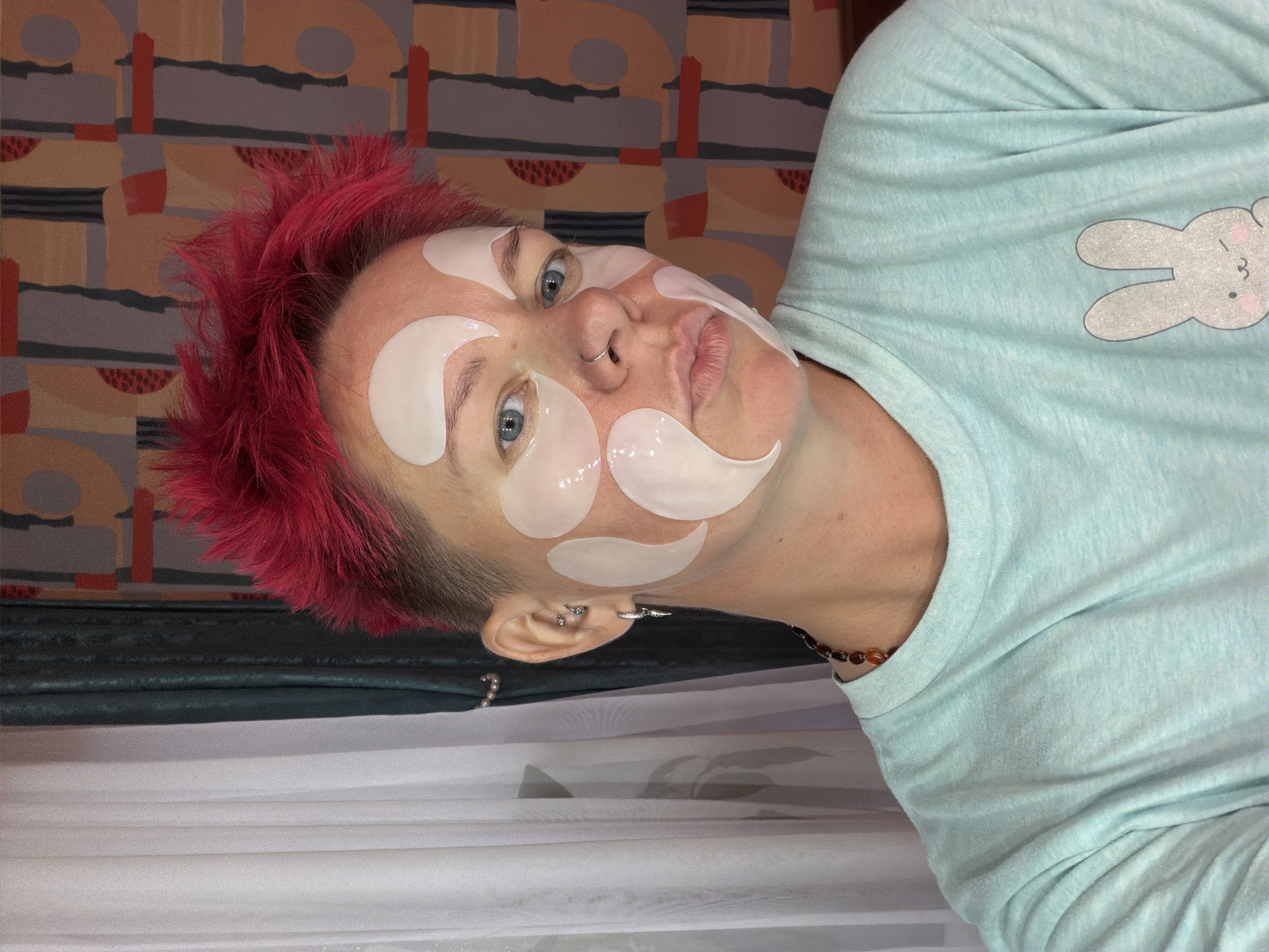 The image shows a person with bright pink, spiky hair and blue eyes. They are wearing a light blue t-shirt with a glittery cartoon bunny printed on it. The person has several white under-eye masks applied to their face. They are also wearing earrings and a necklace. The background appears to be an indoor setting with curtains and a patterned wall. There is no indication of the specific location in the city of Chaikovsky, Russia.