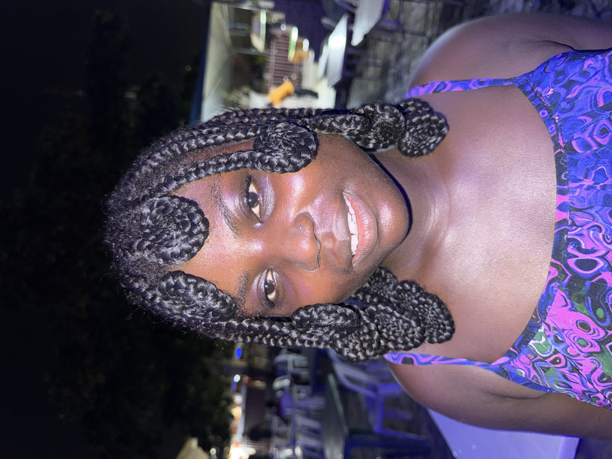 A close-up photograph captures a woman with dark skin in Ado, Nigeria. She is wearing a blue and purple patterned top with thin straps. Her hair is styled in braids, with the ends coiled into buns along her hairline. The background is dark, suggesting nighttime, and includes faint lights and the blurry shapes of outdoor furniture. The focus is primarily on the woman's face, which is slightly tilted toward the camera.