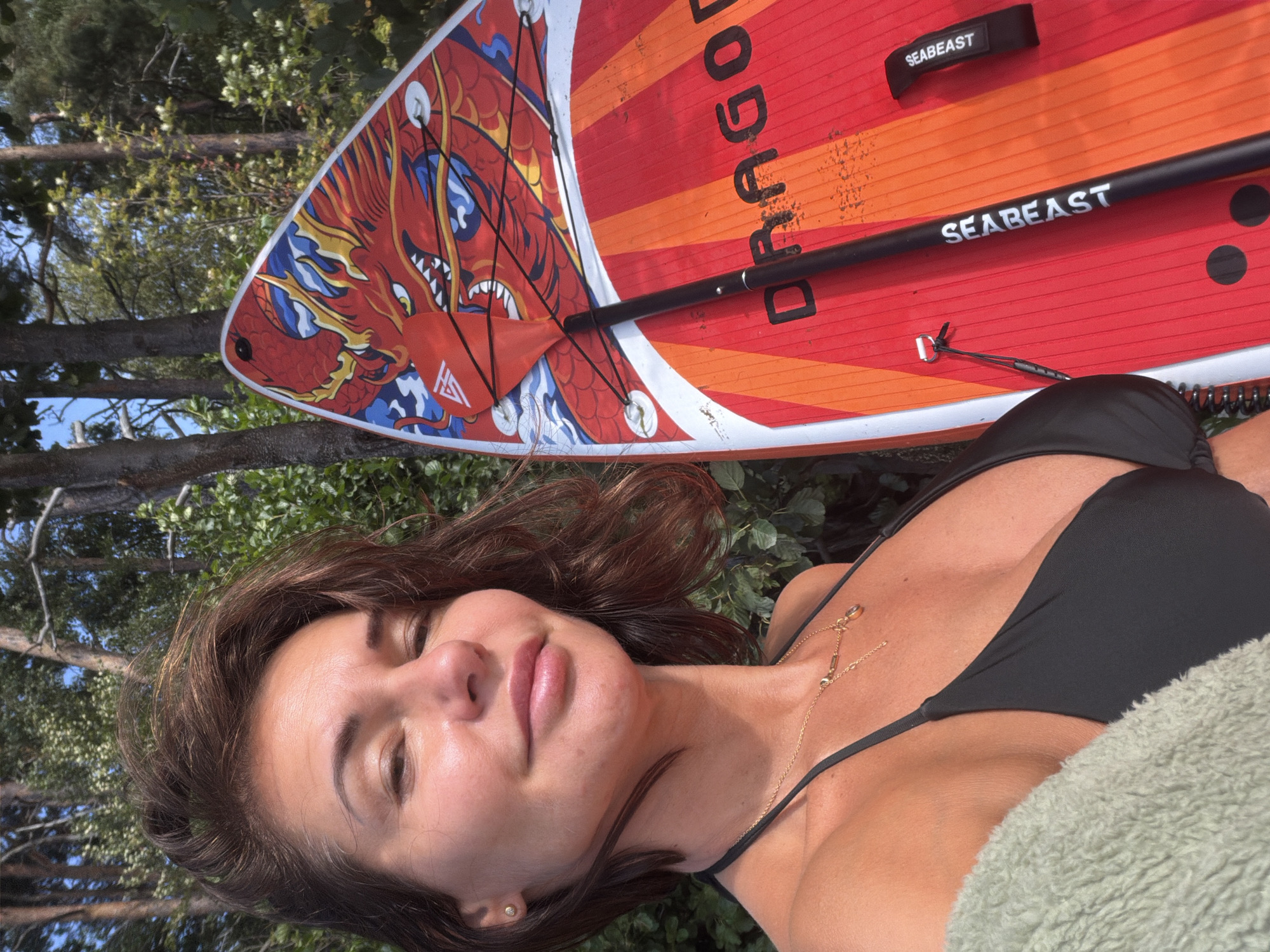 A woman is taking a selfie outdoors, holding a paddleboard and paddle. She is wearing a black bikini top and a light green fuzzy shawl. The paddleboard is red with an orange striped design and a red dragon graphic at the top. The paddle is black with a red blade. The background consists of trees and foliage, suggesting an outdoor setting possibly near a body of water. The woman's face is turned towards the camera, and she is smiling. The image appears to be taken during daylight. There is no indication that the location is in Vyazy, Russia.
