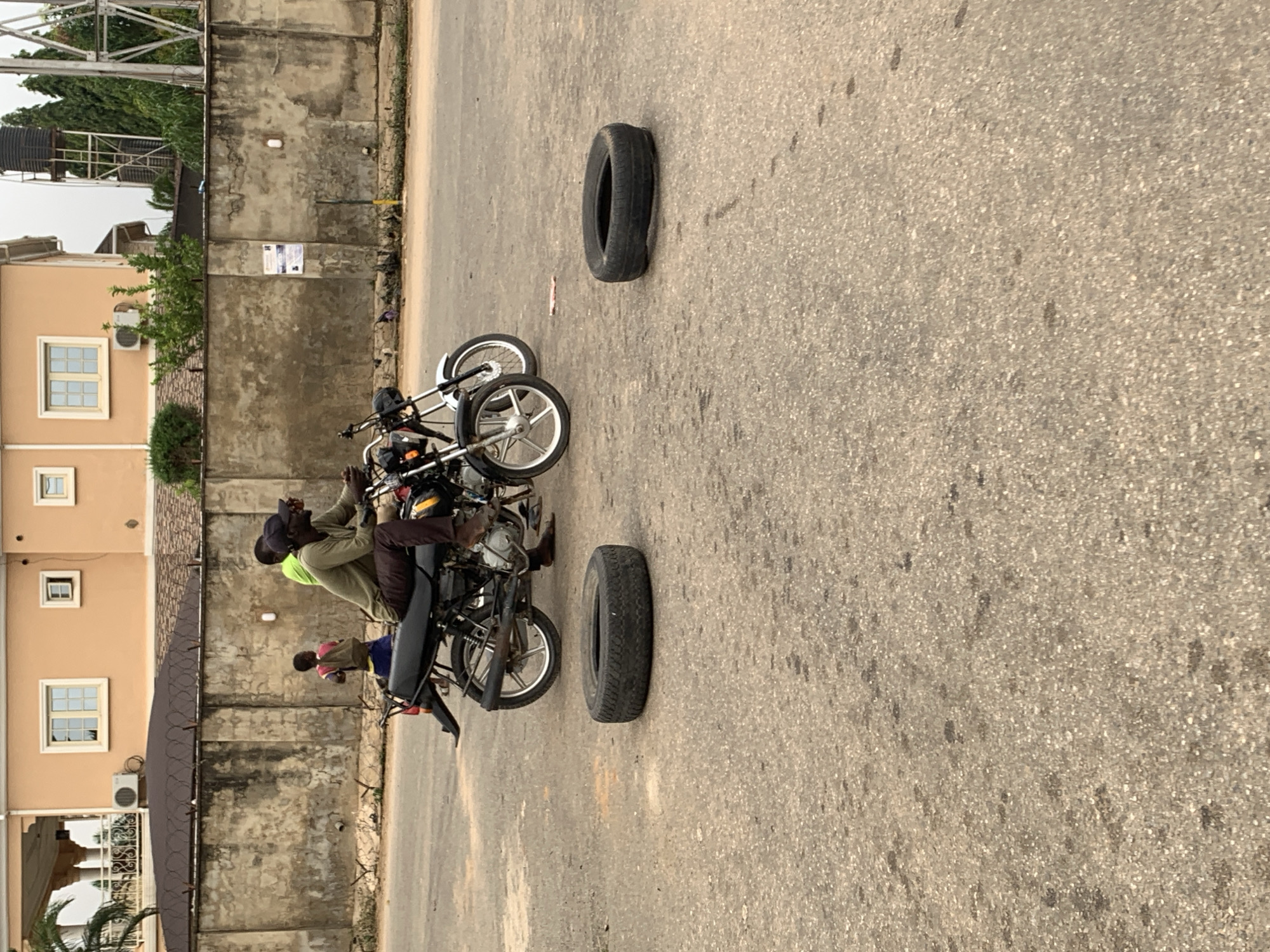 Two men on a motorcycle are parked on a paved road. The motorcycle is black and silver with a black seat. The man in the driver's seat is wearing a dark green long-sleeved shirt and dark pants. The man behind him is wearing a bright green t-shirt and dark pants. Two spare tires are lying on the ground in front of the motorcycle. In the background, there is a beige building with several windows and an air conditioning unit. A concrete wall with barbed wire runs along the property line. A young boy in a pink shirt and blue shorts is standing near the wall. The scene appears to be in an urban setting in Abuja, Nigeria.