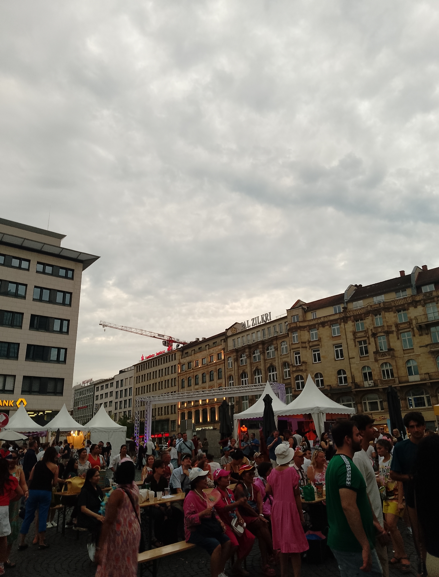 This media file depicts an outdoor gathering of people in a city square. The scene is filled with a diverse crowd, some sitting at long tables, others standing and mingling. White tents are set up, suggesting a market, festival, or outdoor event. People are dressed casually, indicating a relaxed atmosphere, and several appear to be enjoying themselves, with smiles visible on some faces.
In the background, there are multi-story buildings with numerous windows, characteristic of an urban environment. A construction crane is visible above the buildings, and there's some scaffolding. The sky is overcast with grey clouds, suggesting it might be late afternoon or early evening, and the weather is possibly cool or about to rain.
There is visible text on some of the buildings and signage. On the far left, a yellow sign with a black "A" and "NK" is partially visible. Above one of the buildings, text reads "Frankfurter Sparkasse." Further along the buildings, "PAL ZILIERI" is prominently displayed. Another sign reads "ROSSMANN." Some tents also have signage, though it is too blurry to read. The overall impression is a lively city event taking place under a cloudy sky, with a mix of people enjoying food, drinks, and social interaction.