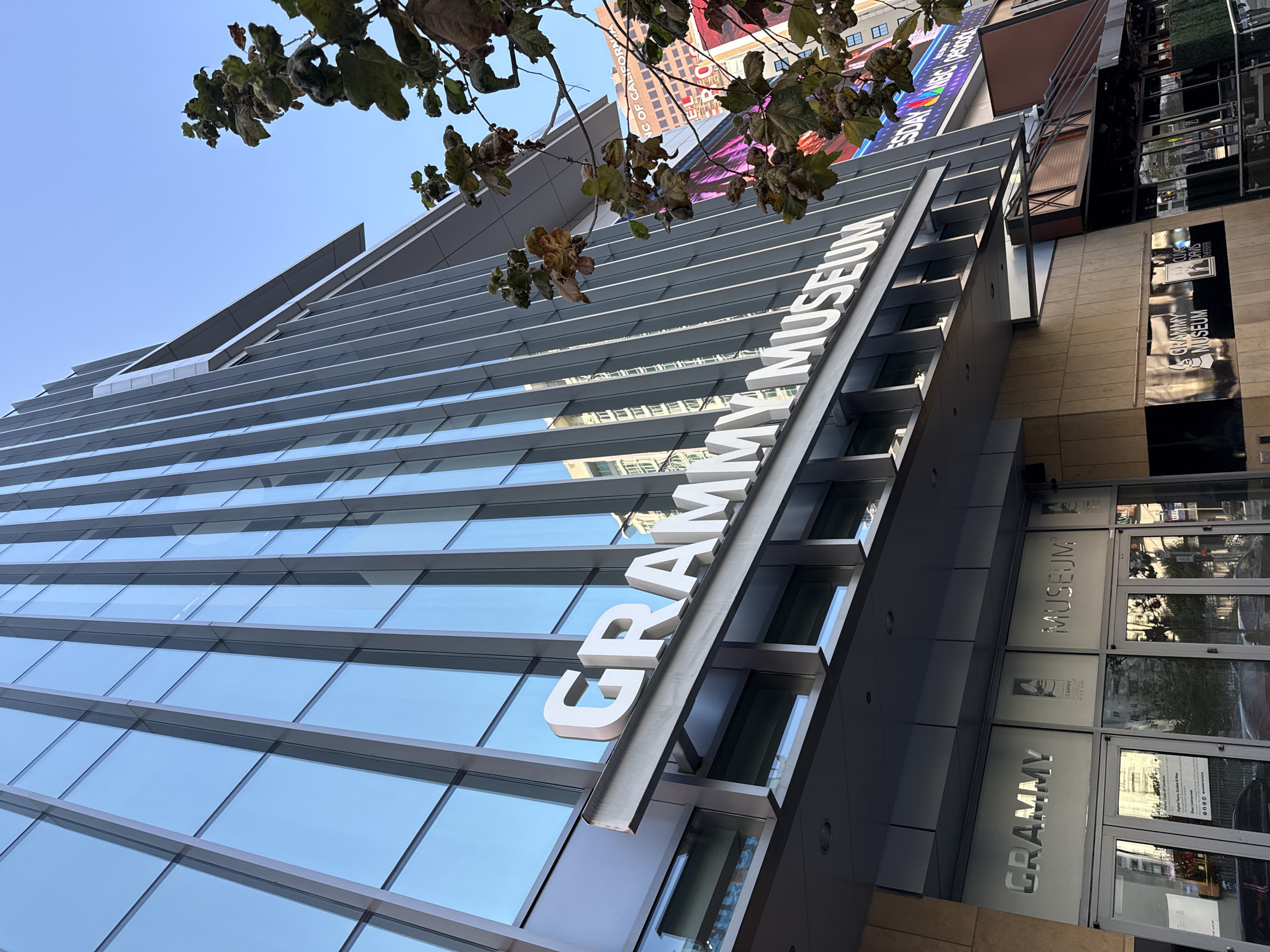 This is a medium shot, low-angle photograph of the Grammy Museum, located at 888 Figueroa Street in downtown Los Angeles, California, United States of America. The building features a modern glass facade with vertical silver accents. Large white three-dimensional letters spelling "GRAMMY MUSEUM" are mounted on a metallic awning above the entrance. Below the awning, frosted glass panels on the building's facade also display "GRAMMY" and "MUSEUM" in large white lettering, with a logo between them. The entrance consists of glass doors and windows, with reflections of the surrounding city visible. A tree with brown, dry leaves branches into the frame from the right side of the image. In the background, other city buildings are visible, including a building with a billboard displaying "NBC". The sky is clear and blue. There are no people or animals visible in the immediate foreground.