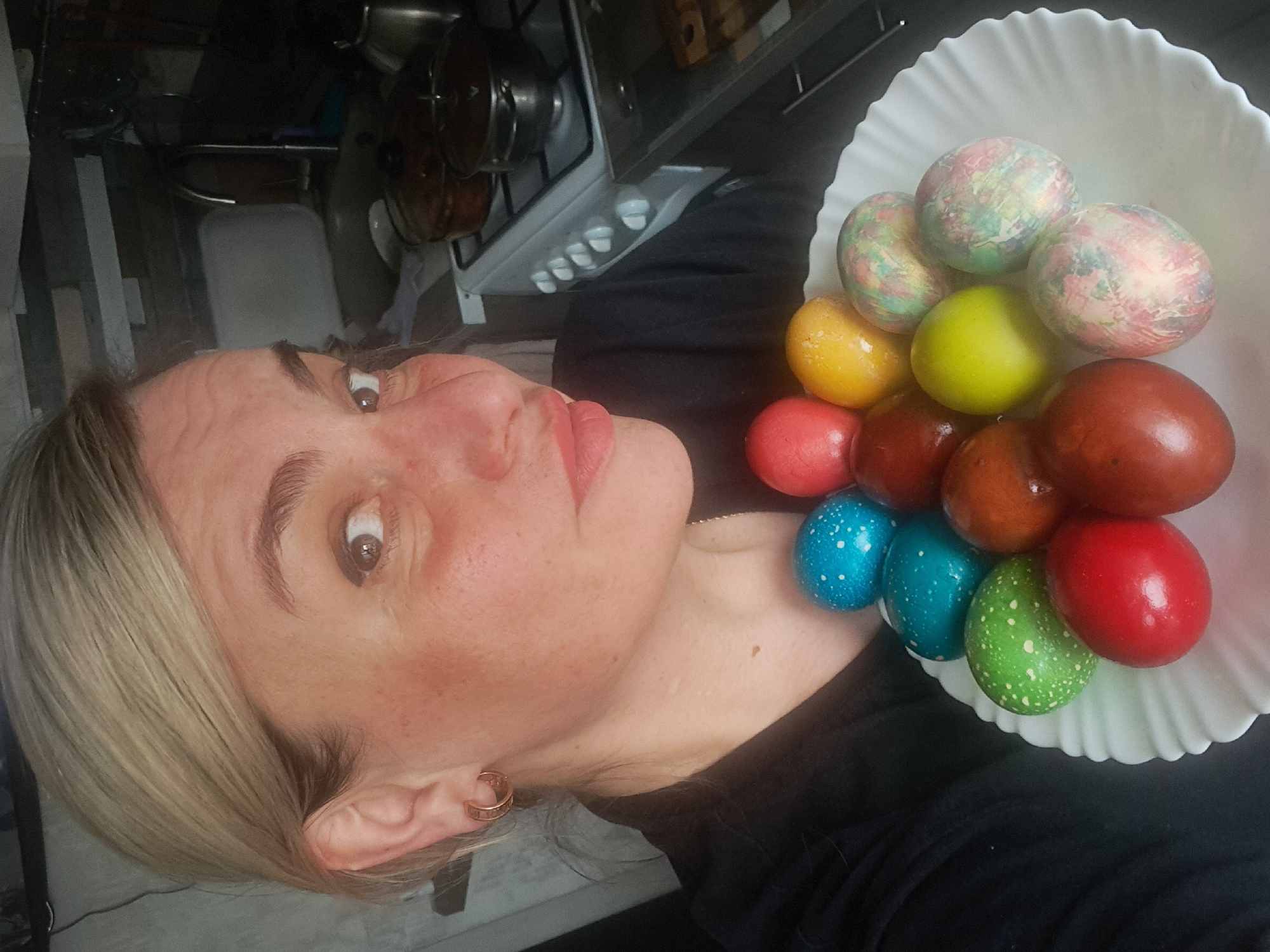 A woman with light blonde hair and light skin holds a white bowl filled with Easter eggs. The eggs are various colors including red, brown, green, yellow, and blue. Some are solid colors while others are decorated with dots or a marbled pattern. The woman is wearing a black shirt. In the background is a kitchen with a white stove and several pots on the stovetop. The location is Elektrostal, Russia.