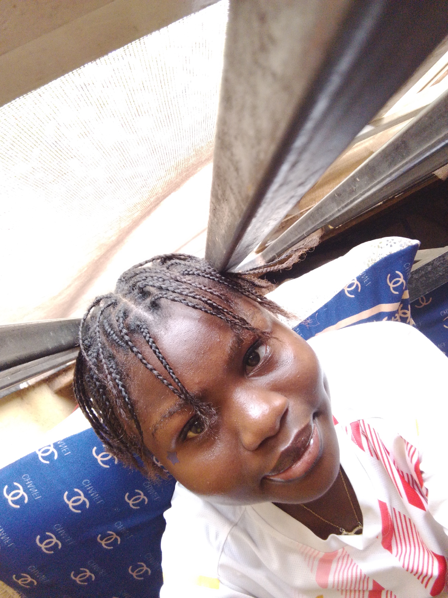 A close-up, high-angle self-portrait shows a young woman with braided hair, seated in what appears to be a bus in Nsukka, Nigeria. She is wearing a white top with red stripes and a delicate gold necklace. The bus interior includes a patterned blue and white seat cushion and a metal window frame above her. A window screen is visible in the background. The light is bright, suggesting daytime. She is smiling slightly.