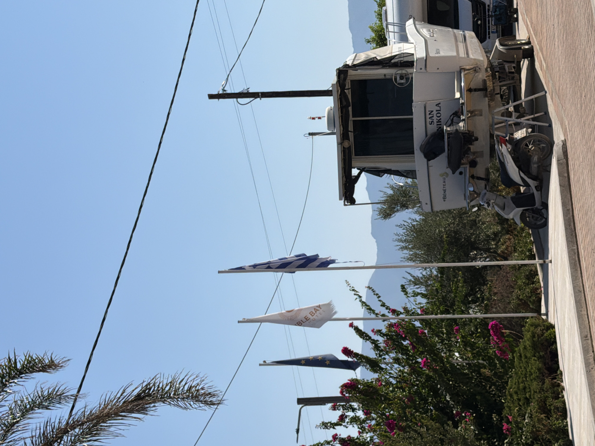 The image shows a boat on a trailer, parked next to a scooter on a paved area. There are three flags flying on poles: the European Union flag, a white flag with orange lettering that appears to say "BUNGALOW HOTEL," and the Greek flag. Lush green bushes with pink flowers are visible to the left of the flags. In the background, there are mountains and a clear blue sky. Electrical wires are strung across the upper portion of the image. The boat has the name "SAN NIKOLA" written on its stern. A white vehicle, possibly an RV, is partially visible to the right of the boat. The scene appears to be in an outdoor setting in a sunny climate.