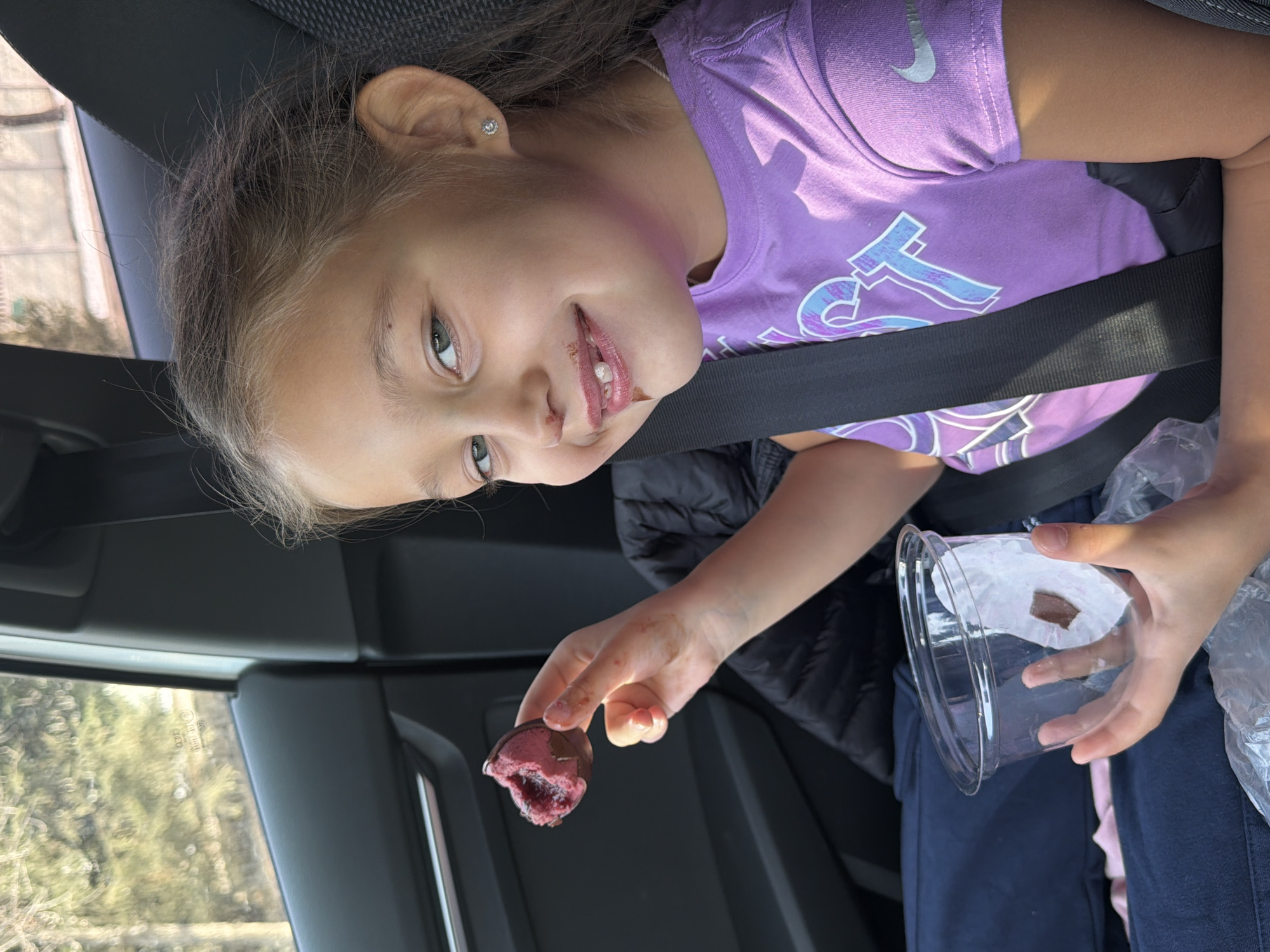 A young girl is depicted inside a car, smiling cheerfully towards the camera while holding a partially eaten dessert. She has light brown hair and green eyes, with food residue visible around her mouth, indicating she is in the process of eating. She wears a vibrant purple t-shirt featuring a silver "Nike" swoosh logo on the sleeve and a larger, stylized light blue and white graphic, possibly "FT", across the chest. A black seatbelt is securely fastened over her, suggesting she is seated in a moving vehicle, likely the back seat.
In her right hand, the girl holds a chocolate-covered treat with a prominent pink or reddish filling. Her left hand holds an empty, clear plastic cup, presumably the original container for the dessert, next to a crumpled plastic bag. The car's interior is visible, and daylight filters through the window, illuminating a blurry background of trees outside, indicating it is daytime. Faint text, including "E43" and "DOT", is discernible on the car window glass. The girl's happy expression suggests she is enjoying her snack. This scene is set in Krasnoyarsk, Russia.