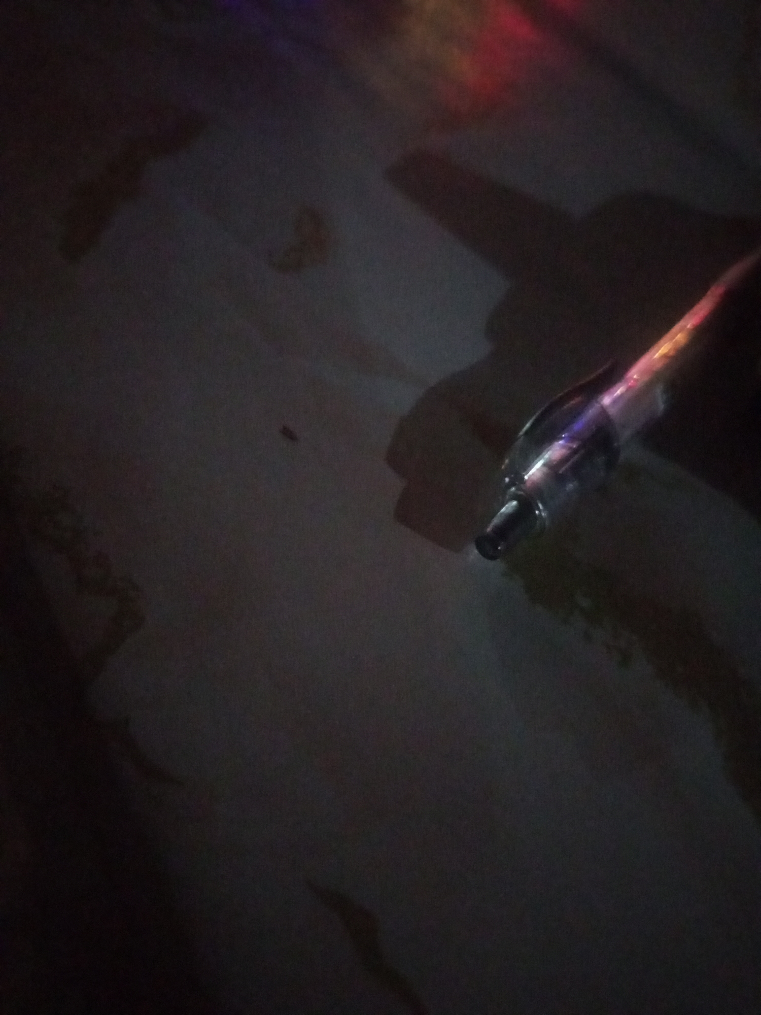 The image shows a close-up, dimly lit view of a dark surface, possibly a table or floor, with a slight sheen. A small, dark, insect-like object is visible in the mid-left of the frame. In the right half of the image, a thin, transparent tube, possibly a light stick or similar object, is angled from the upper right towards the lower left. The tube is illuminated from within, casting colorful light that reflects off the dark surface. The background is mostly dark with abstract shapes of reflected light in shades of red, pink, and purple visible in the upper portion. There is no indication of the specific location within Lhokseumawe, Indonesia, nor any people or distinct interactions.
