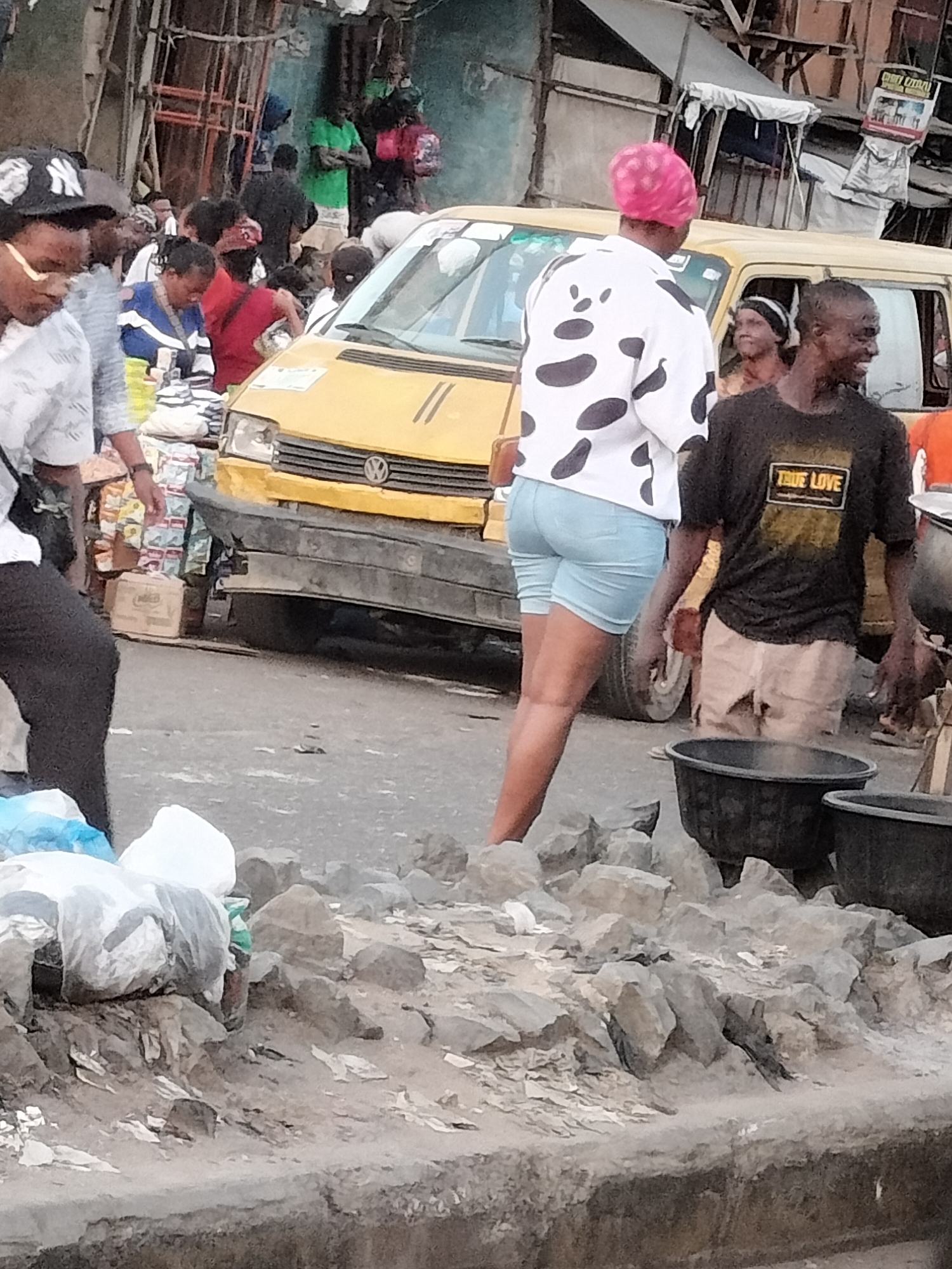 The image depicts a bustling street scene in Lagos, Nigeria. A yellow Volkswagen van is partially visible, surrounded by pedestrians. A woman wearing a spotted white shirt and pink headscarf stands in the foreground. Beside her, a man in a "TRUE LOVE" T-shirt carries two large black buckets. The foreground is filled with piles of gray rocks and debris. In the background, other people are shopping, and a man with a baseball cap is on the left. The environment is characteristic of a busy urban market area in Lagos.