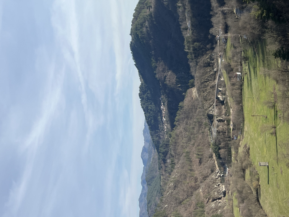 The image depicts a panoramic view of a mountainous valley, situated in the vicinity of Stoianovo, Bulgaria. The upper half of the frame shows a light blue sky with wispy white cirrus clouds, indicative of a clear day.
In the foreground, the lower third of the image is dominated by vibrant green fields, interspersed with scattered bare deciduous trees and their shadows. Two distinct structures are visible in these fields: a dark, rectangular, vertical utility box or small shed, and a taller, slender metal lattice structure, likely an electricity pylon.
The midground features a winding river or large stream, its banks lined with sparse vegetation and rocky outcrops. A prominent stone arch bridge, possessing at least three visible arches, spans this waterway, carrying a paved road that curves sharply on both approaches. Beyond the river and bridge, the terrain rises into hillsides covered predominantly by bare deciduous trees and some evergreen conifers, suggesting late autumn or early spring. A small cluster of houses with reddish roofs, representing the village of Stoianovo, is visible nestled on a distant hillside in the mid-right portion of the midground.
The background consists of multiple layers of progressively darker, forested mountains extending to the horizon. No discernible individuals, specific actions, or interactions are visible within the frame. The overall environment is rural and mountainous, illuminated by natural daylight casting distinct shadows.