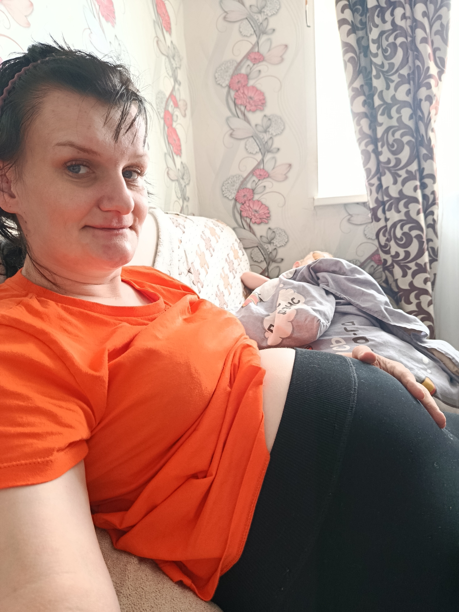 The image shows a woman in an orange t-shirt and black leggings resting on a patterned sofa. A child is lying next to her, covered with a blanket. The background includes a patterned wall and curtains. The location is Фоки, Russia.