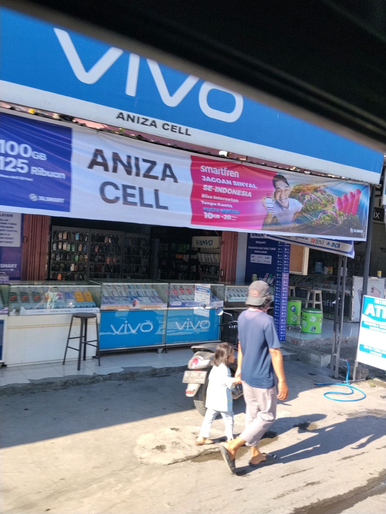 The image shows a street scene in Terara, Indonesia, focusing on a Vivo mobile phone shop called "Aniza Cell". A large blue and white banner with the Vivo logo and "ANIZA CELL" is prominently displayed above the shop entrance. To the right of this banner, a smaller red banner advertises "smartfren JAGOAN SINYAL SE-INDONESIA" with an image of a smiling man holding a phone and a vibrant landscape.
The shop itself appears to be a retail establishment with shelves displaying numerous mobile phones. Below the shelves, glass display cases showcase more phones and accessories. A blue sign with "vivo" printed in white is visible on a counter or display unit.
In the foreground, a man wearing a dark blue t-shirt, grey pants, and a baseball cap is walking away from the camera, accompanied by a young girl in a white top and light-colored pants. They are walking on a paved sidewalk. A motorcycle is parked to the left of the frame, partially obscuring the lower part of the shop. In the background, various shops and advertisements line the street, indicating a commercial area. The lighting suggests it is daytime, with strong sunlight casting distinct shadows on the ground.